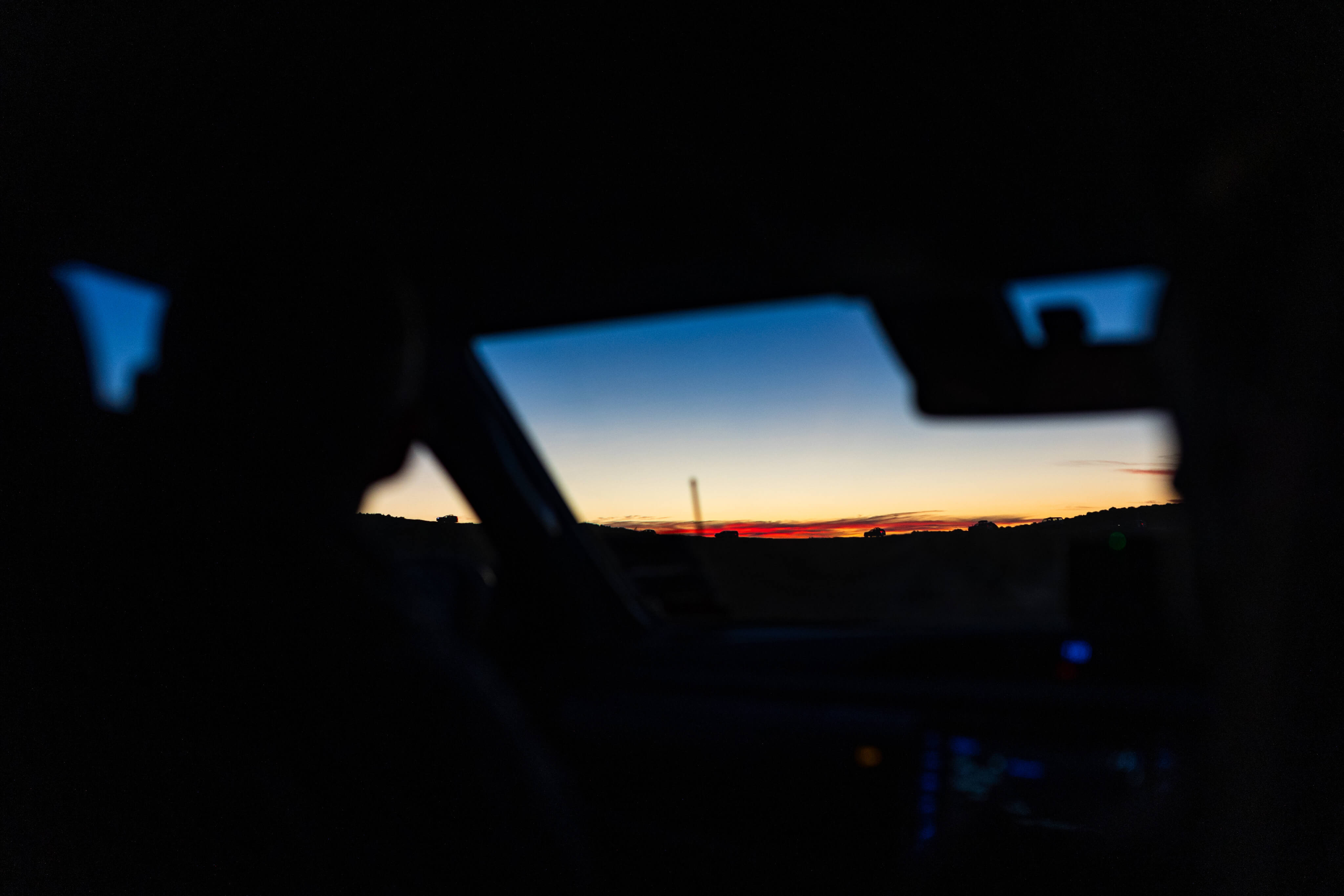 Silhouettes of two people inside a car looking at a colorful sunset through the windshield.