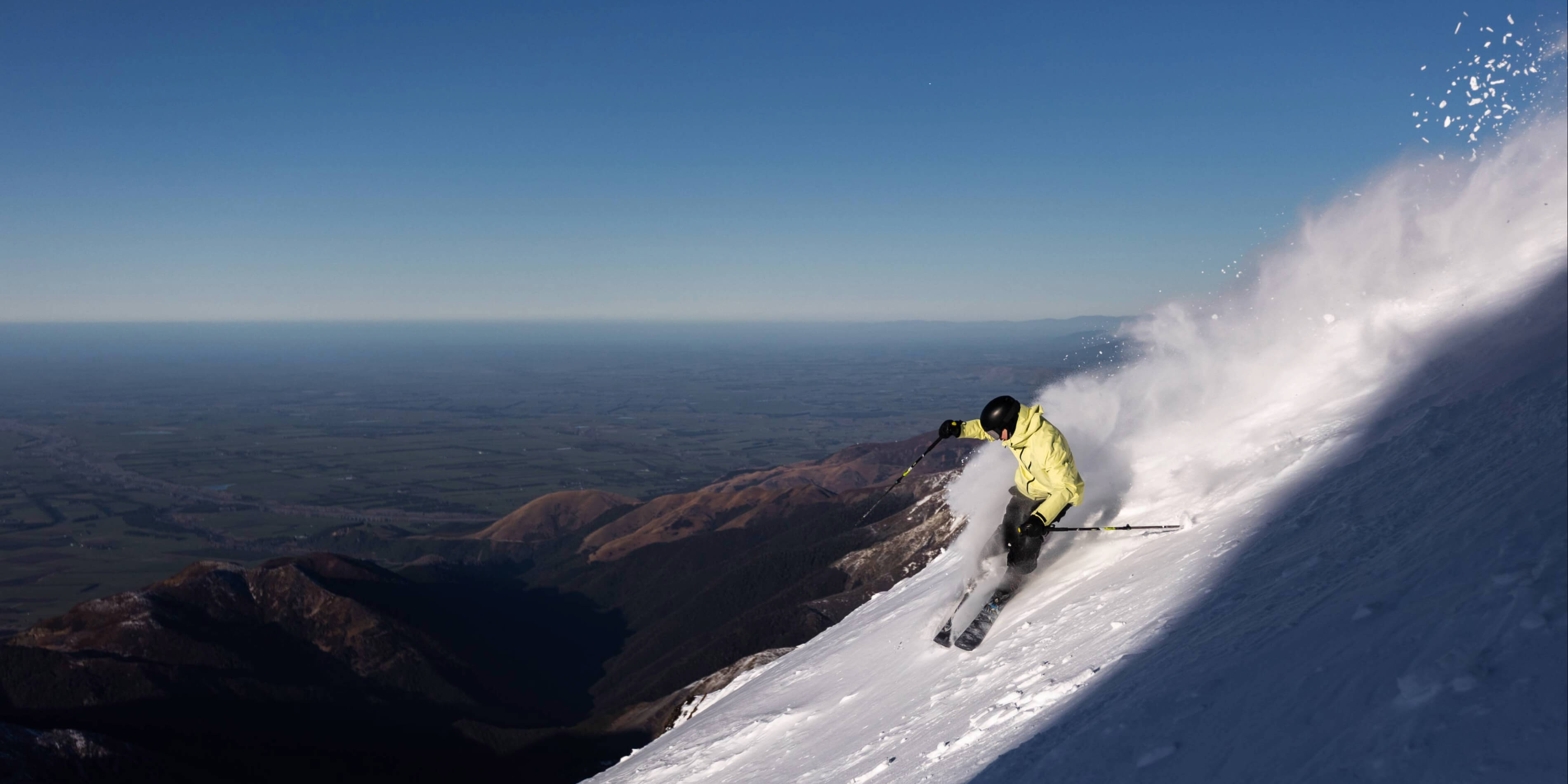 Skier in a yellow jacket skiing downhill on a snowy mountain with powder snow spray and a valley landscape in the background.