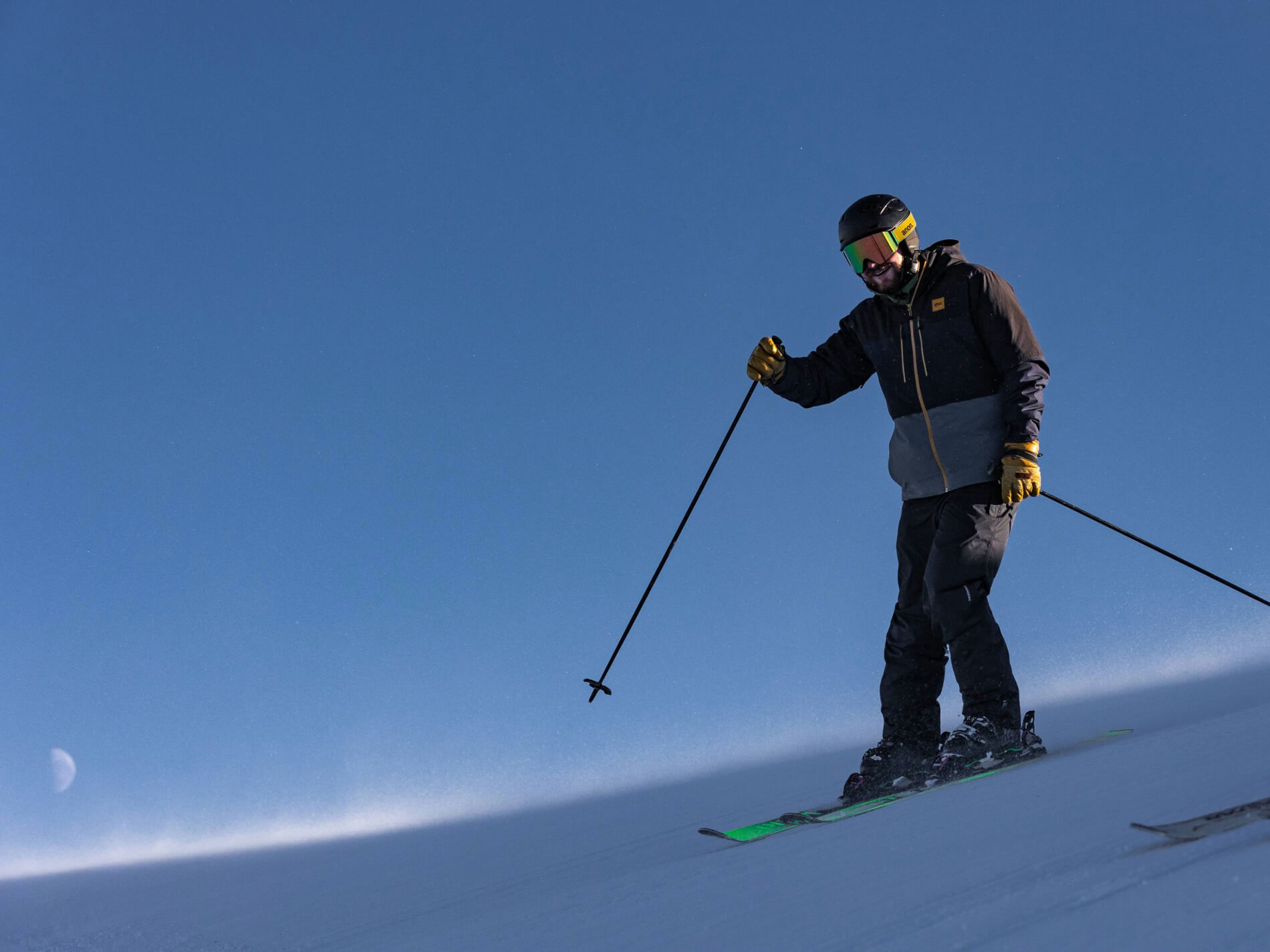 Skier wearing a black and gray jacket, black helmet, and yellow gloves skiing downhill under a clear blue sky with a visible crescent moon.