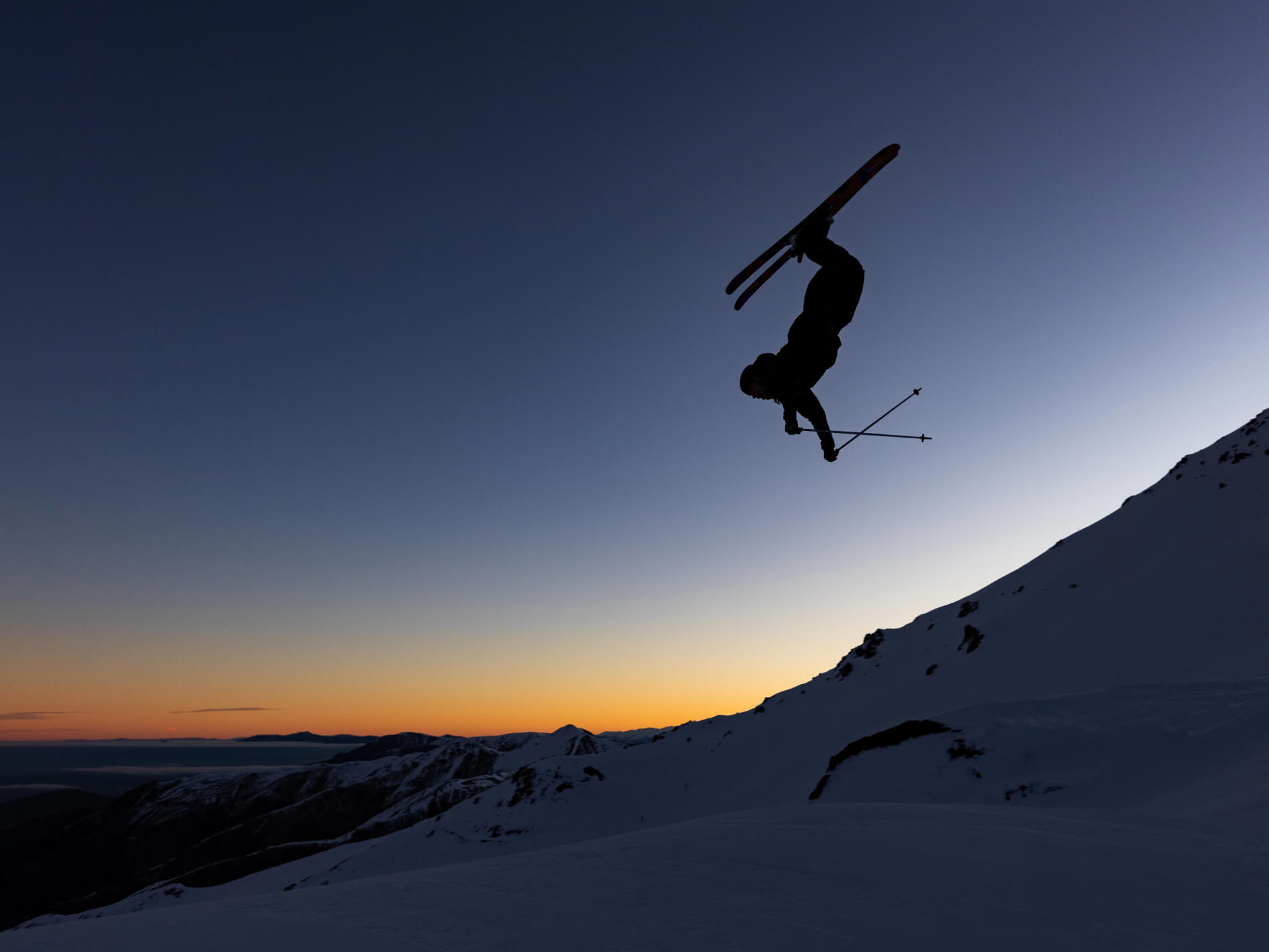 Silhouette of a skier performing a backflip in mid-air over snowy mountains at sunset.