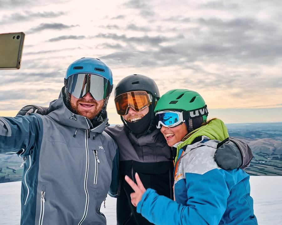 Three skiers wearing helmets and goggles smiling and taking a selfie on a snowy mountain with a cloudy sky in the background.