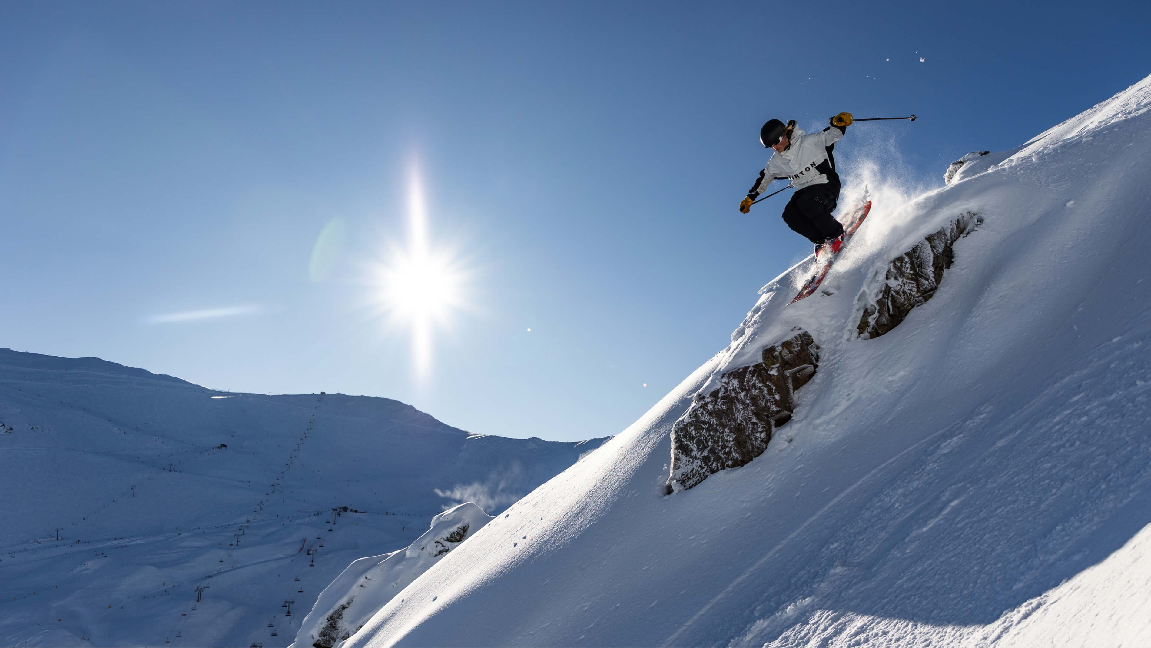 Skier in black helmet and white jacket skiing off a rocky snowy slope with bright sun and clear blue sky.