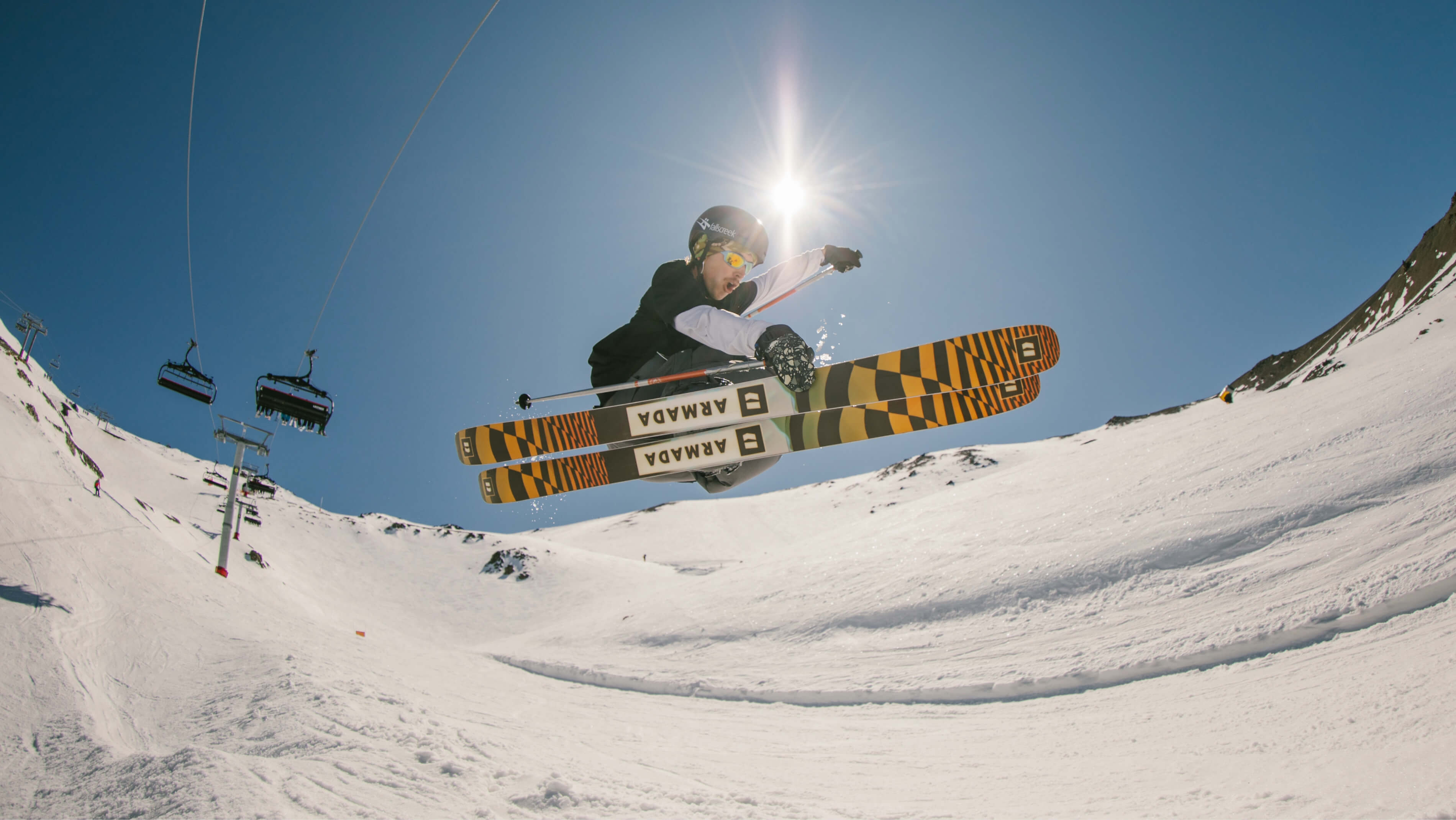 Skier mid-air performing a jump on a sunny snowy mountain slope with ski lifts in the background.