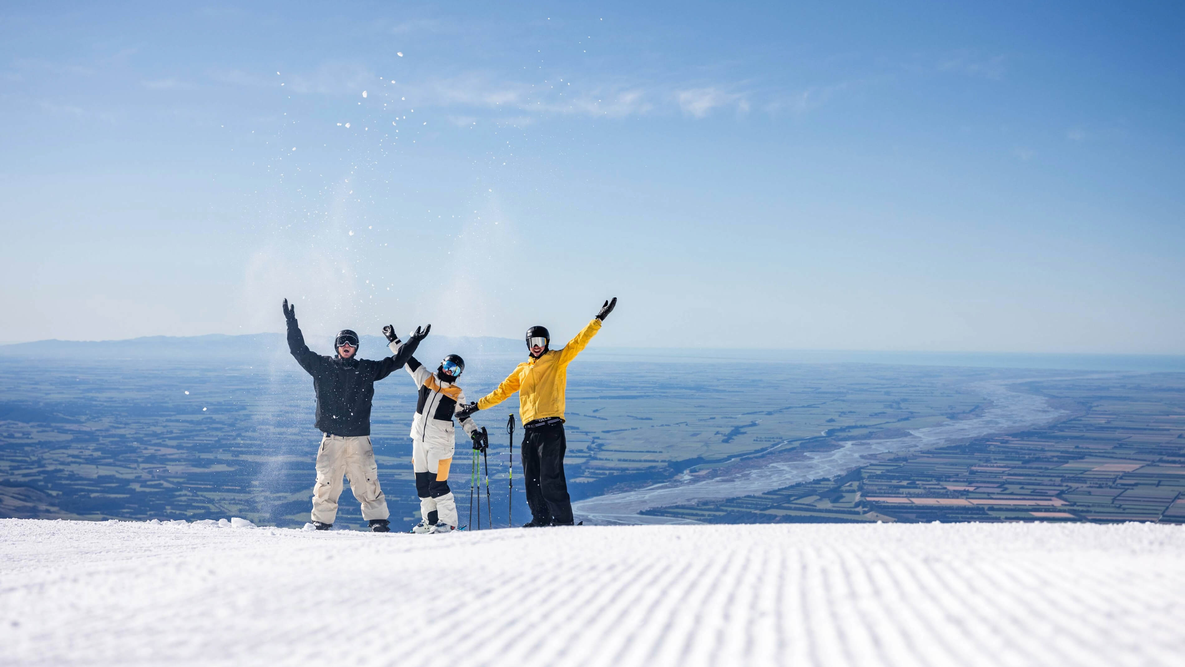 Three skiers in colorful gear celebrating with arms raised on a snowy slope overlooking a vast landscape under a clear blue sky.