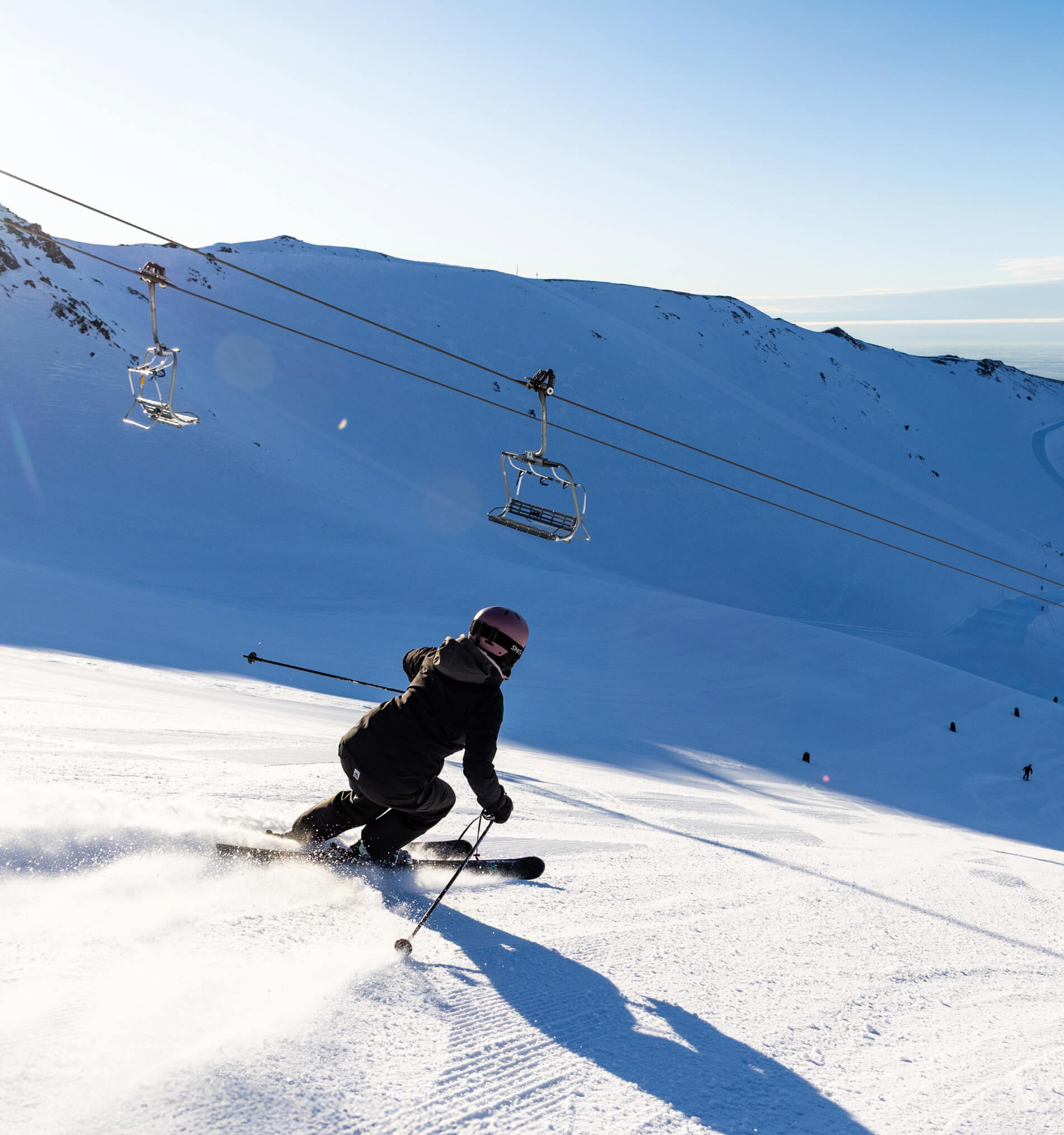Skier wearing a helmet and black jacket skiing downhill on a snowy mountain with ski lift chairs overhead.
