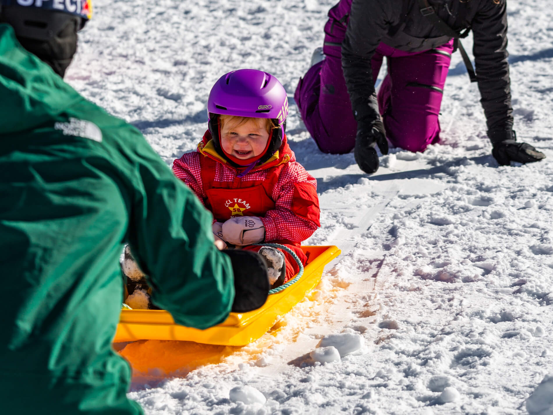 Smiling child wearing a purple helmet and red winter clothes sitting on a yellow sled in the snow.