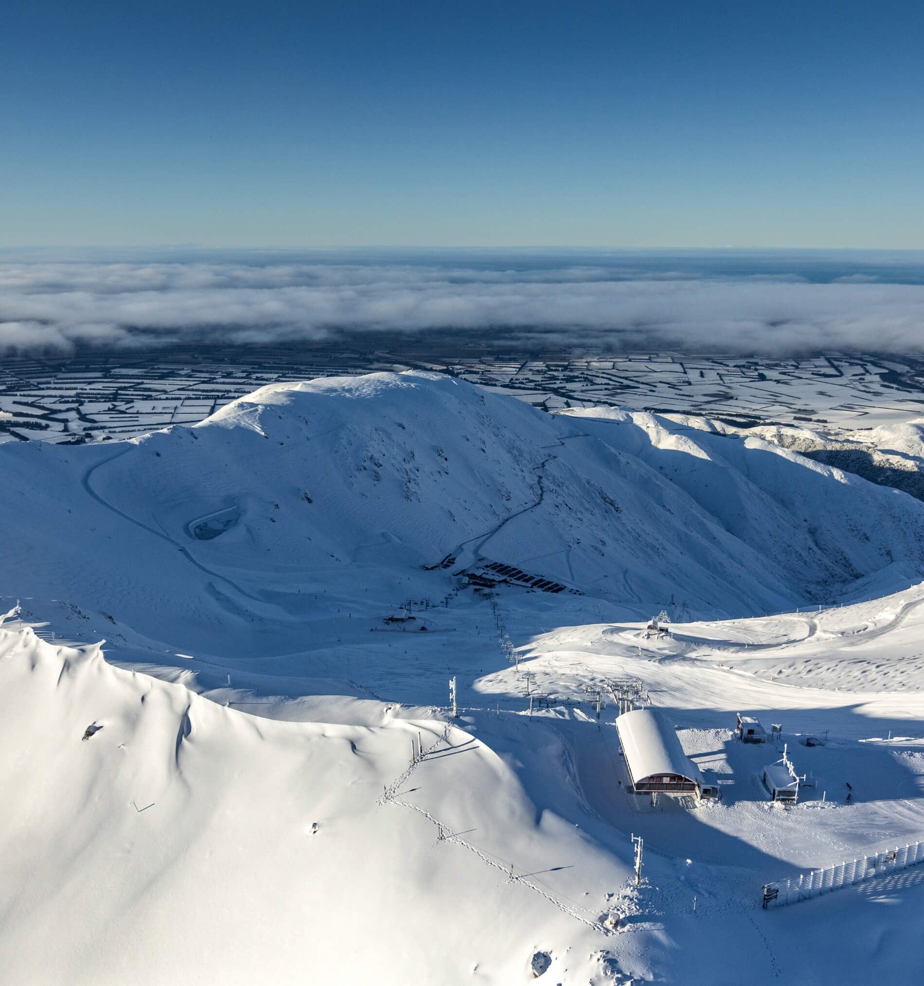 Aerial view of a snow-covered mountain with ski lift structures and tracks, under a clear blue sky and fog over farmland in the distance.