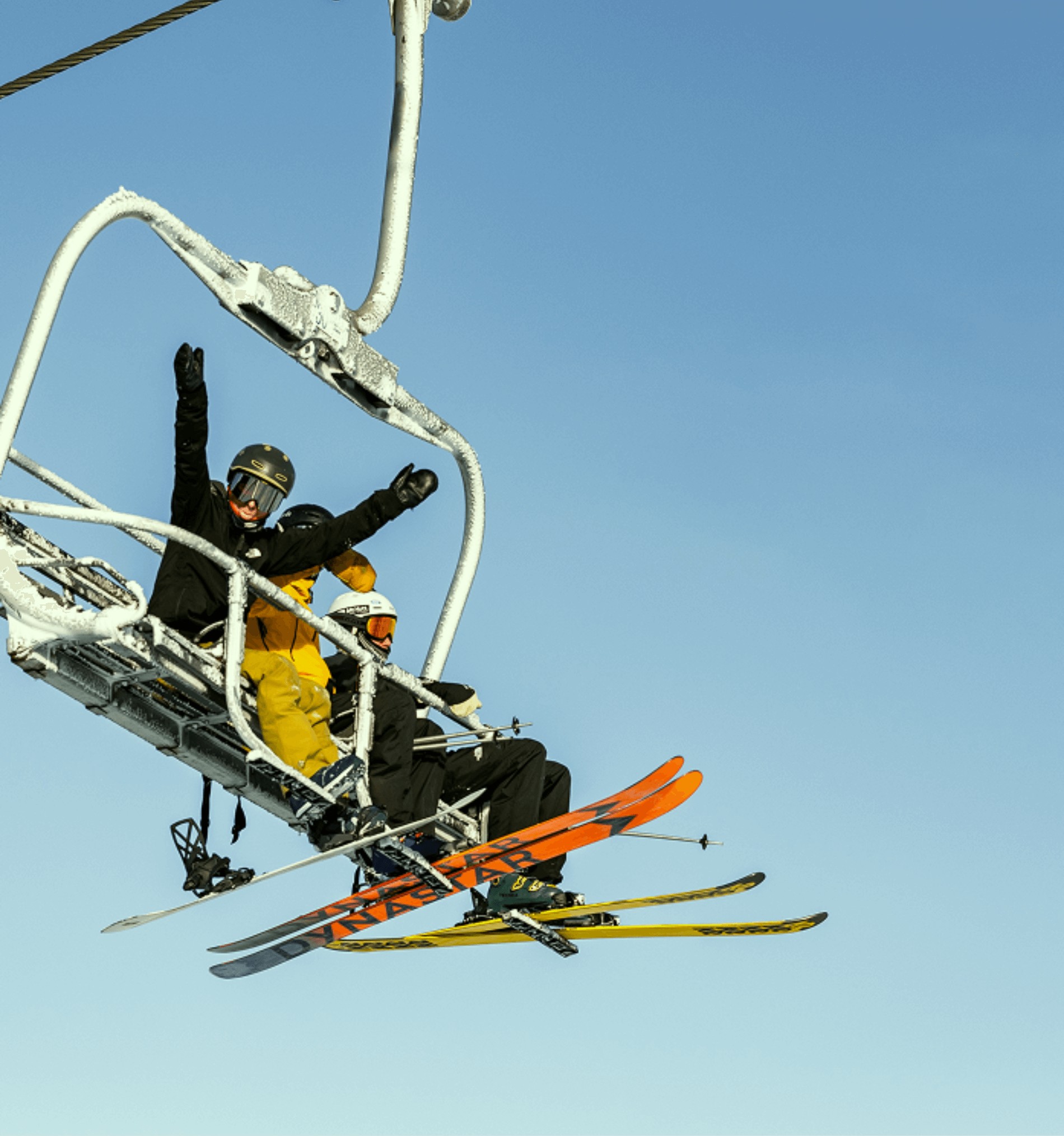 Three skiers sitting on a ski lift against a clear blue sky, two are waving and wearing helmets and ski gear.