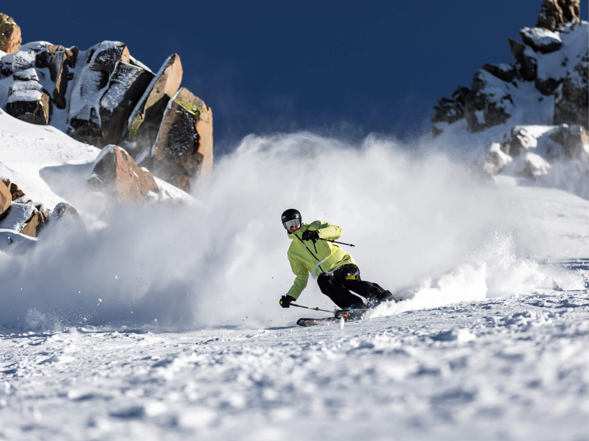 Skier in a bright yellow jacket carving through snow on a mountain slope with rocky outcrops under a clear blue sky.