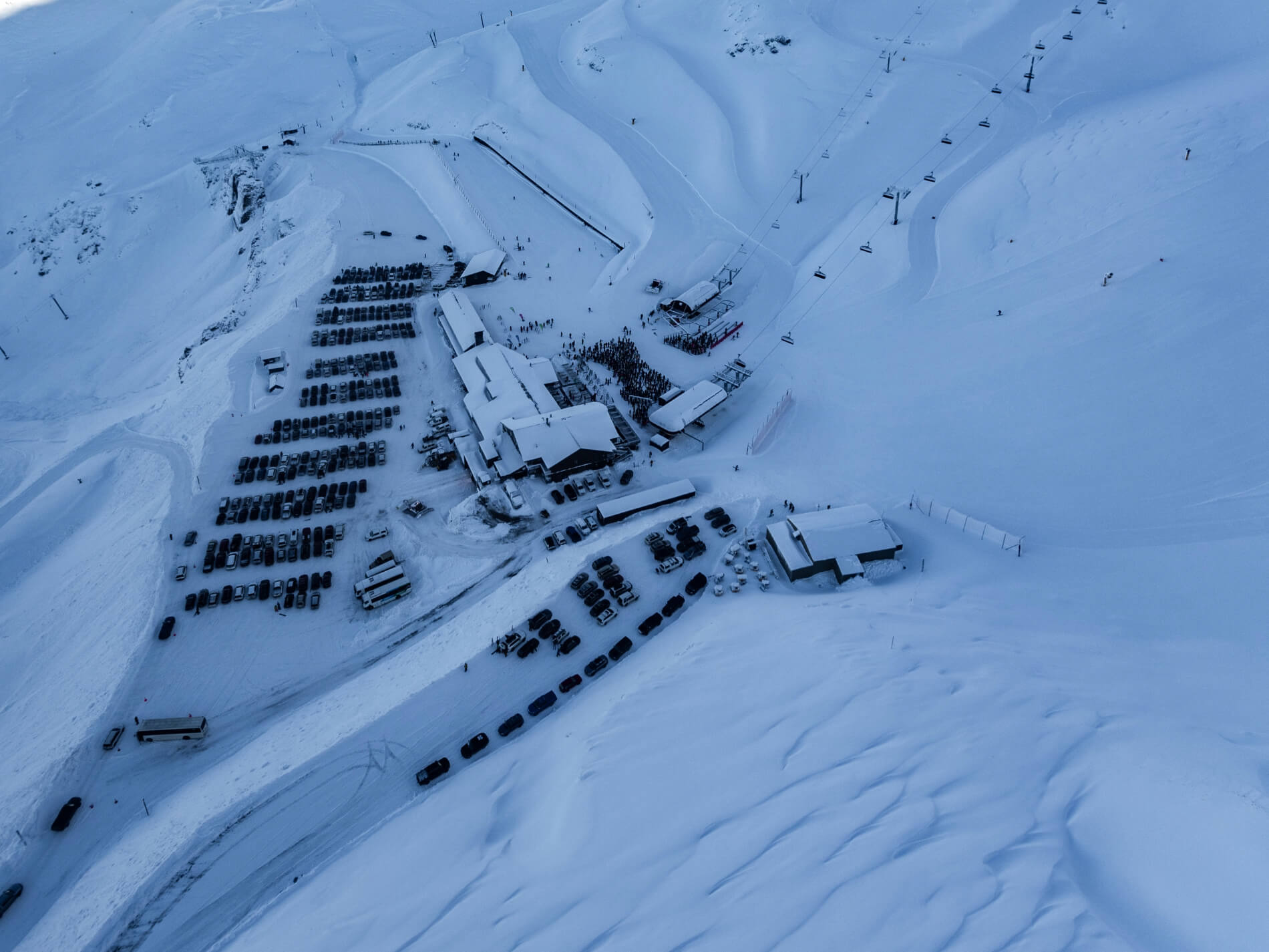 Aerial view Mt of Hutt Ski area covered in snow. Featuring the parking lot with cars, buildings, ski lift, and skiers on slopes.