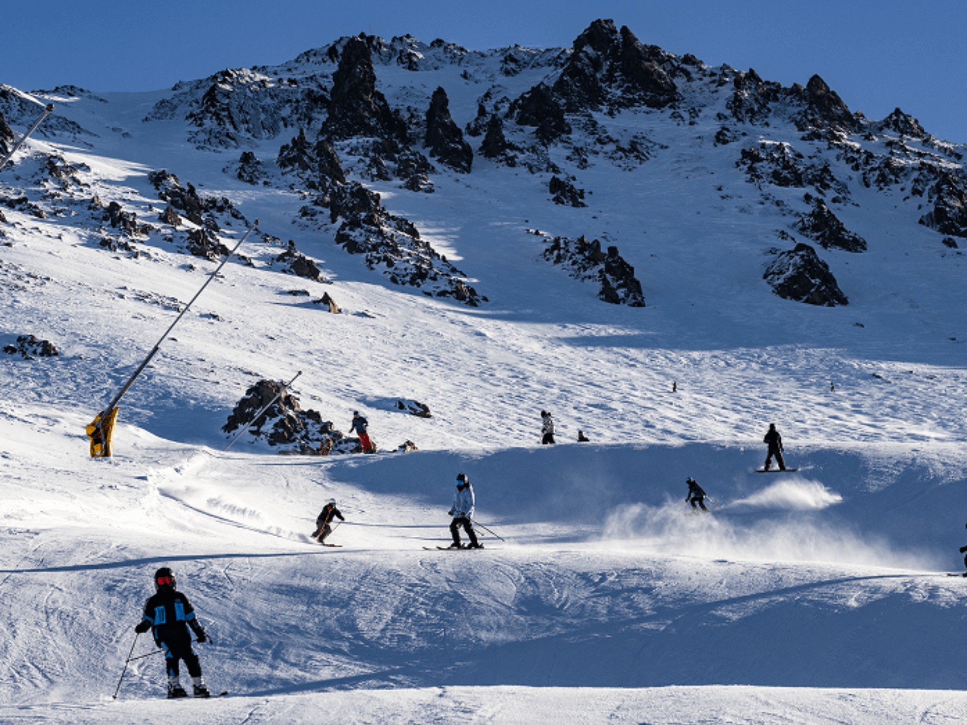 Skiers and snowboarders on a snowy mountain slope with rocky peaks under a clear blue sky.