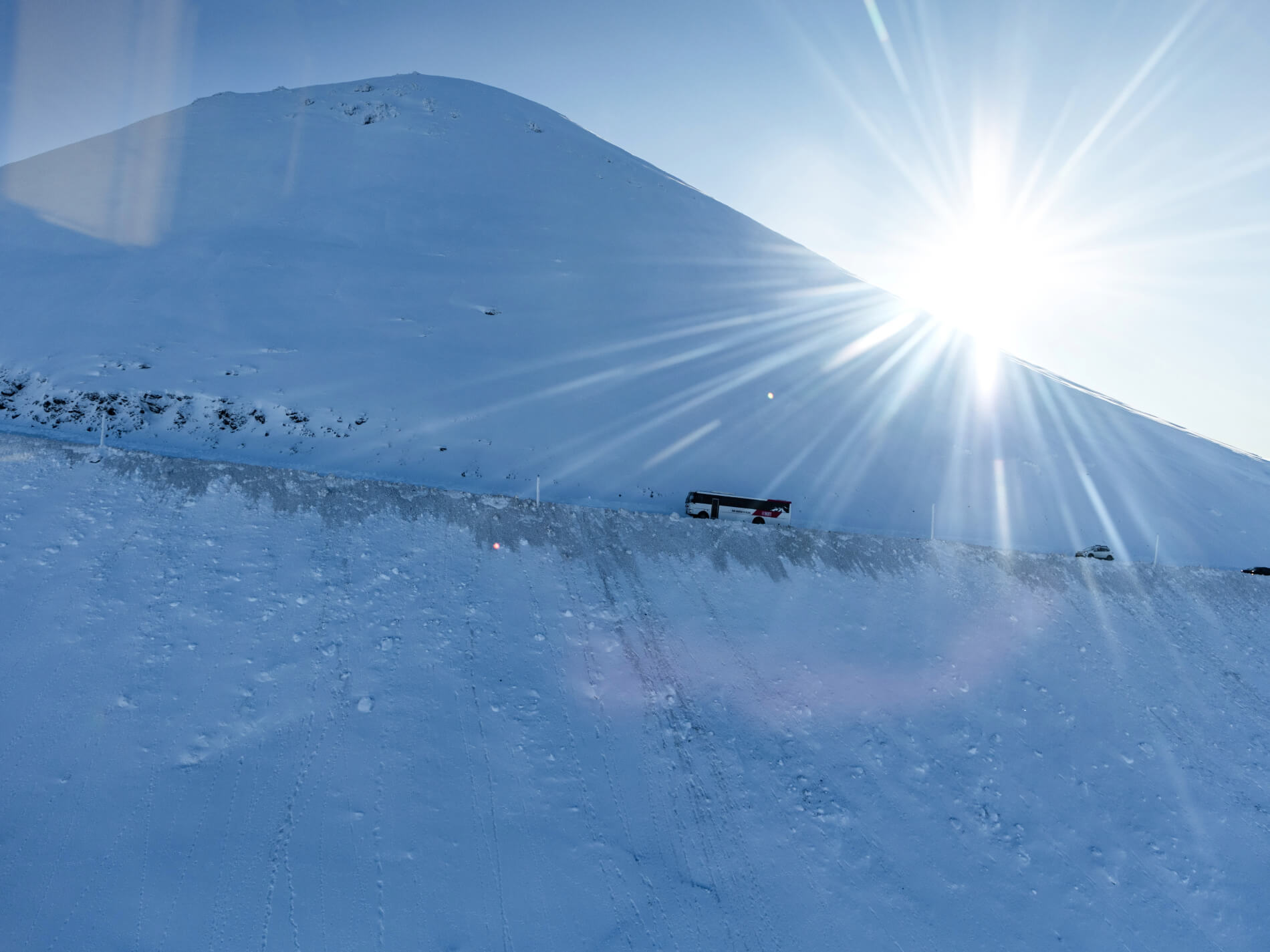 Sun shining over a snowy mountain with a road where a bus and cars are traveling.