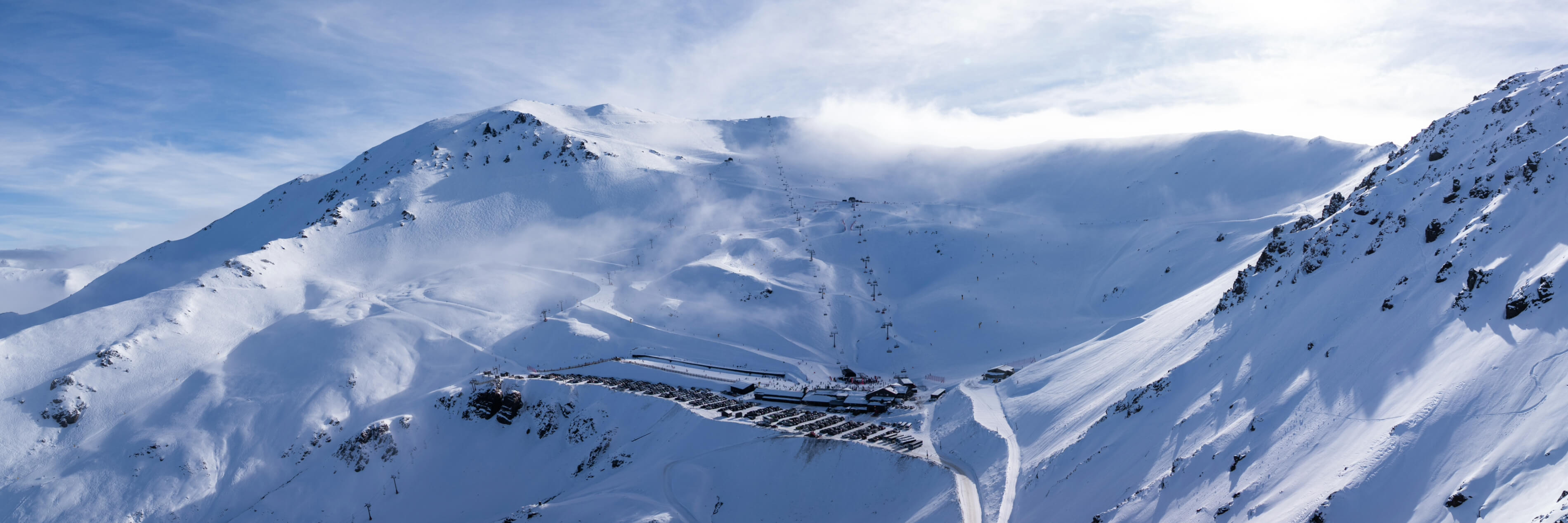 Snow-covered ski resort in a mountain valley with ski lifts, winding snowy trails, and parked cars visible at the base.