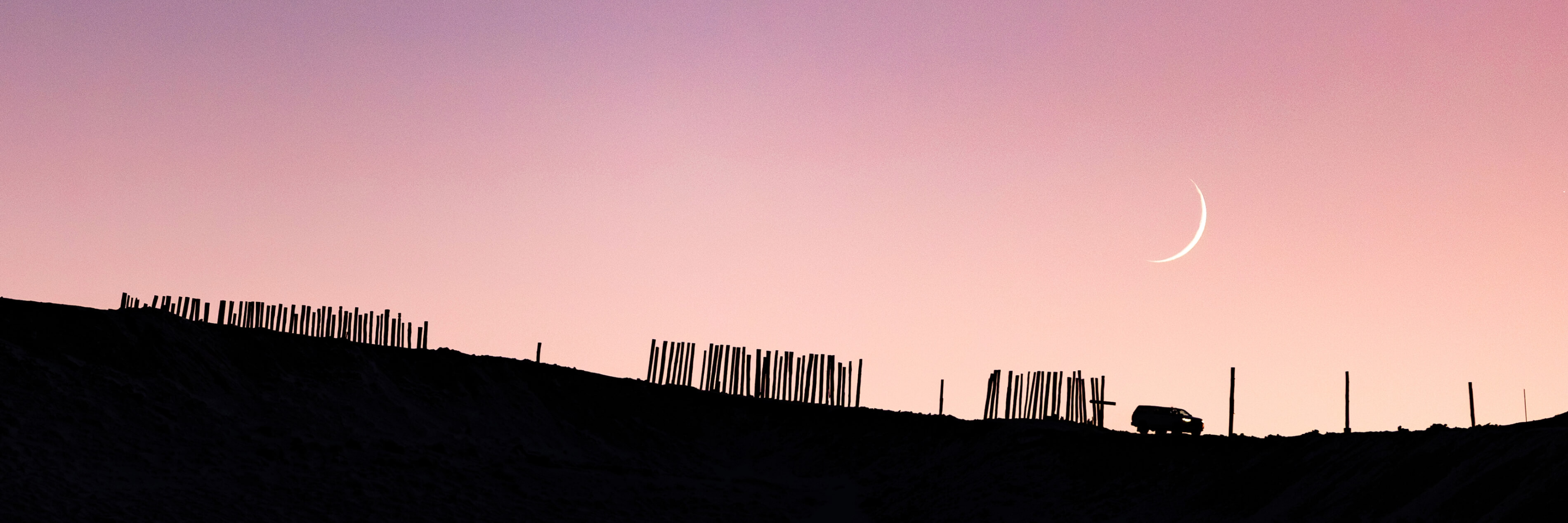 Silhouetted fence posts and a vehicle on a ridge under a crescent moon in a pink twilight sky.