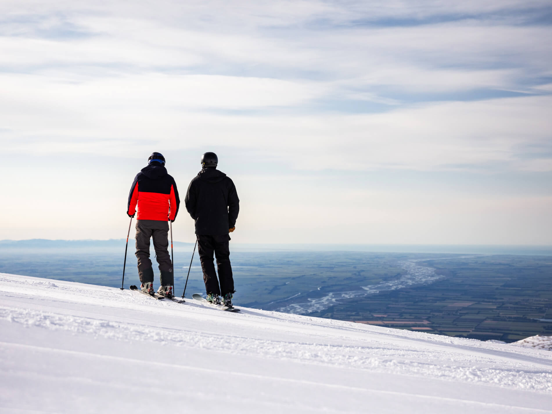 Two skiers standing on a snowy slope overlooking a vast landscape with fields and a river under a partly cloudy sky.