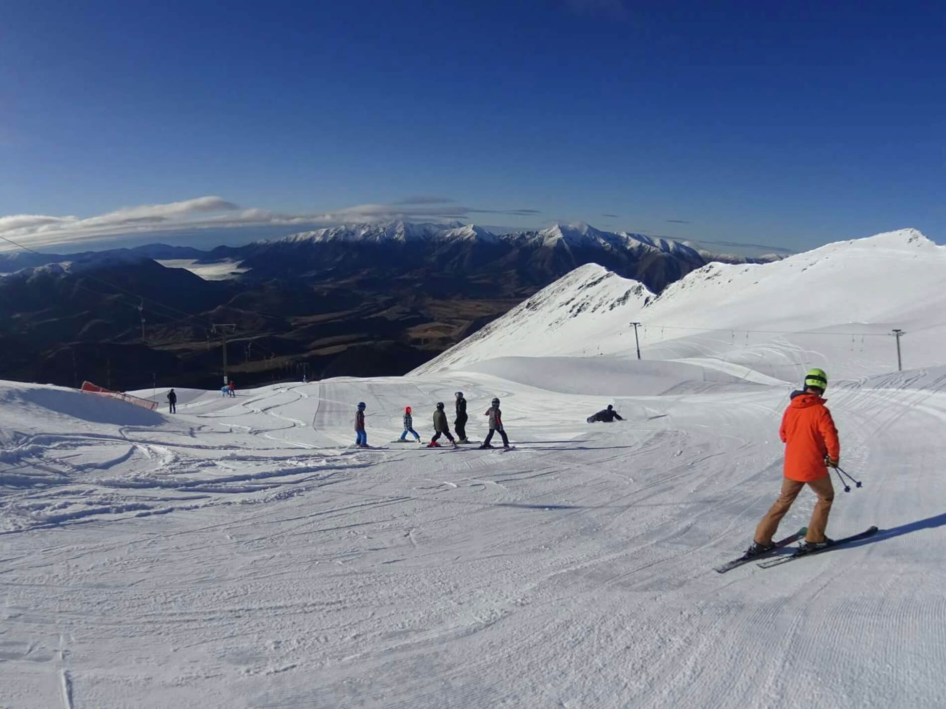 Group of skiers on a snowy mountain slope with snow-covered peaks and clear blue sky in the background.
