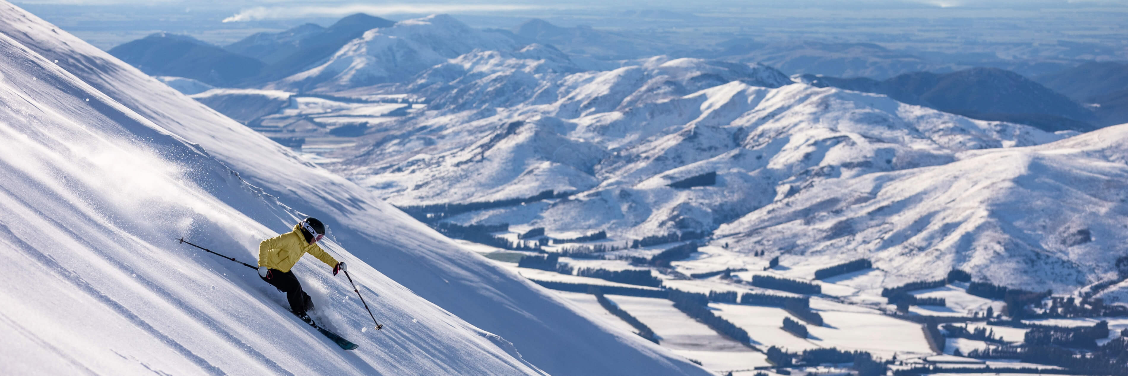 Skier in a yellow jacket skiing down a snowy mountain slope with snow-covered hills and a valley in the background.