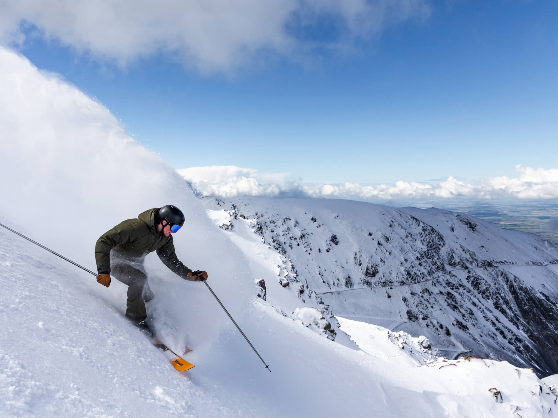 Skier in dark clothing and helmet carving through deep powder snow on a steep mountain slope under a blue sky.