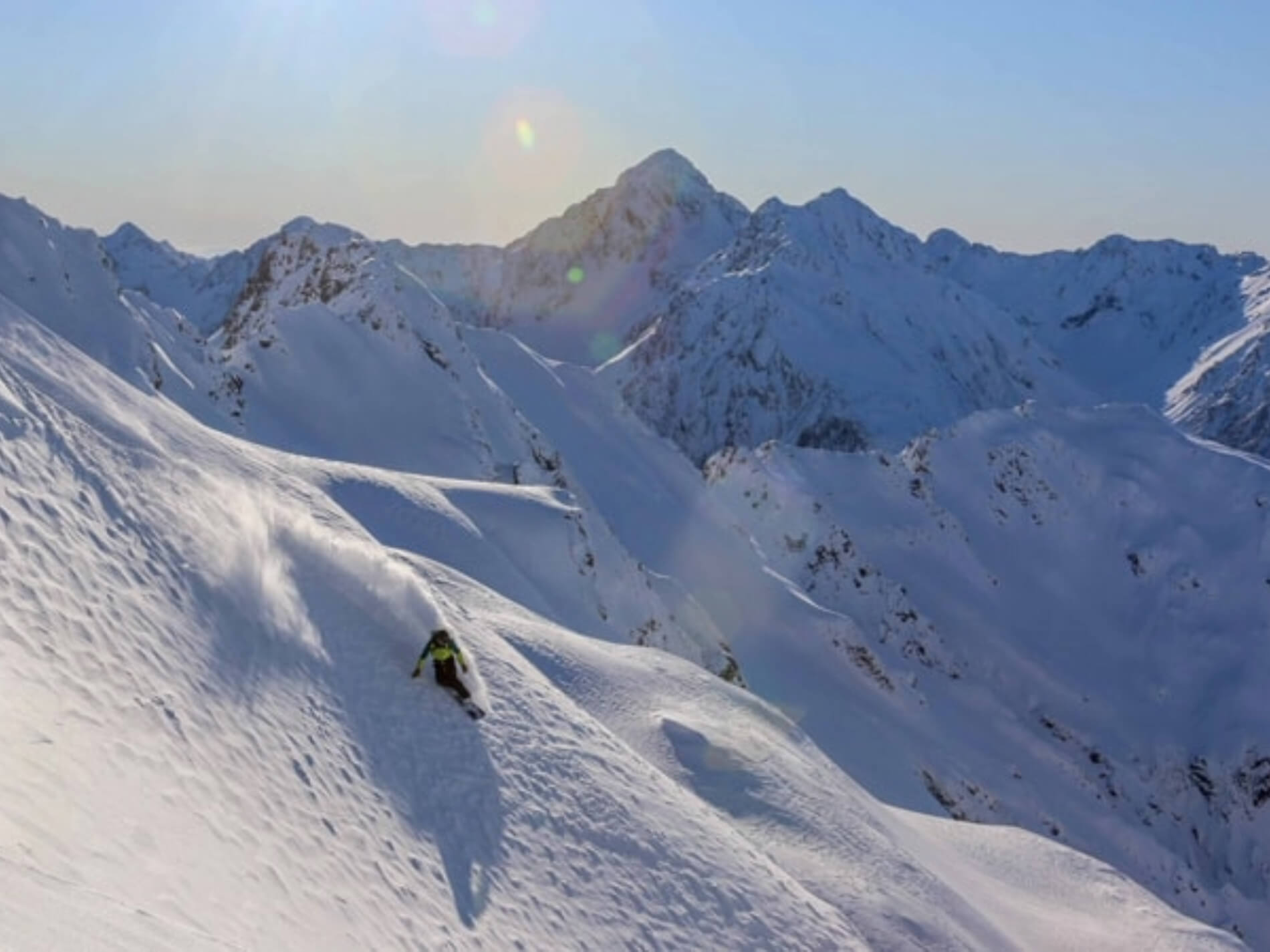 Snowboarder carving down a snowy mountain slope with sunlit peaks in the background.