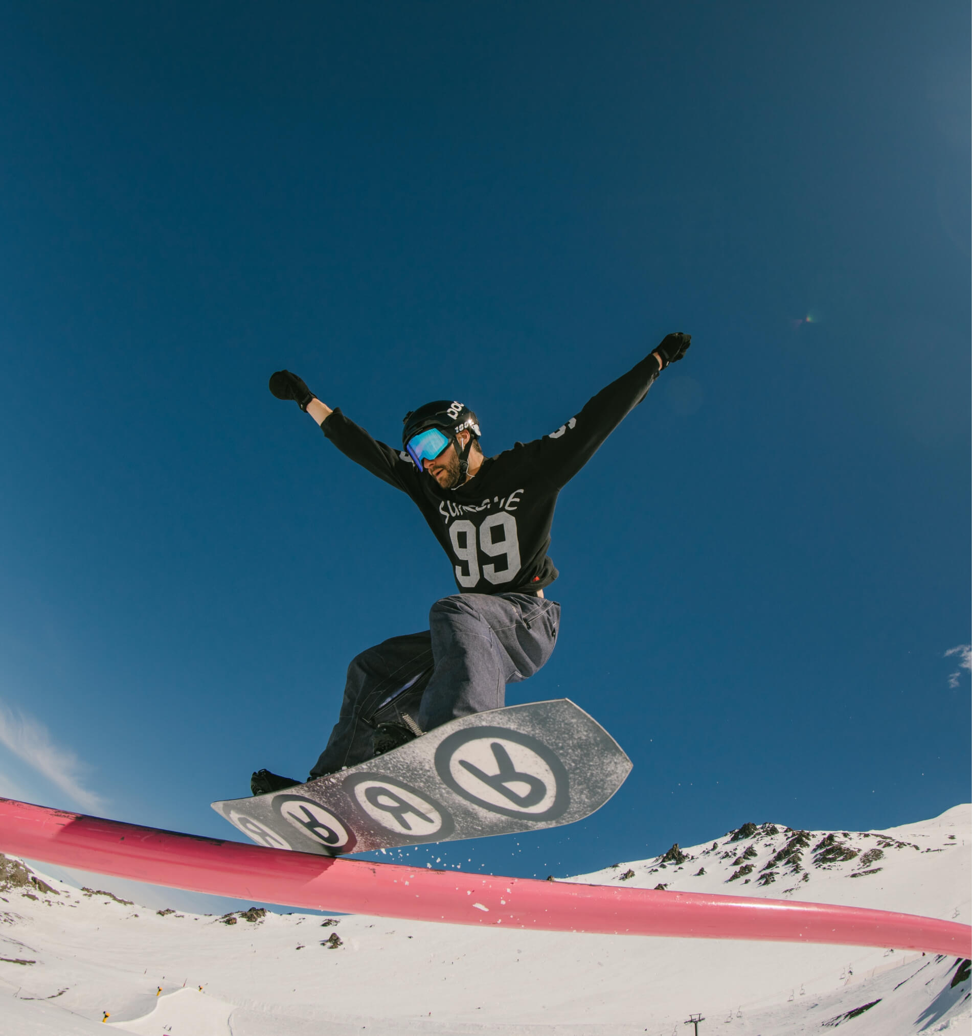 Snowboarder in black helmet and goggles jumping over pink rail on snowy mountain under clear blue sky.