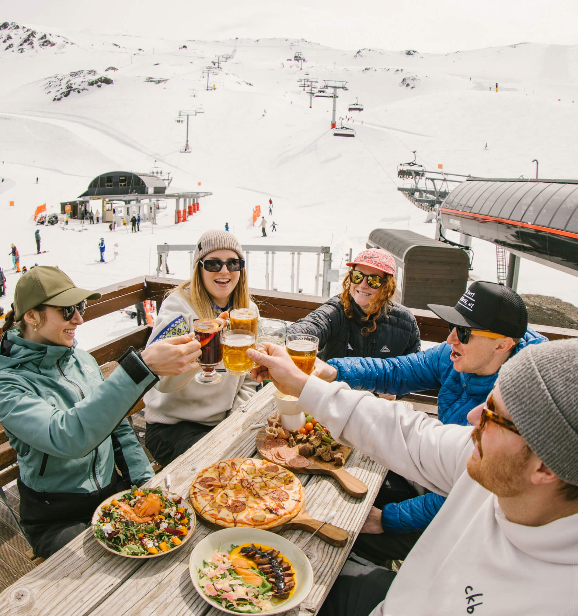 Five friends wearing winter clothing toast with drinks around a picnic table with pizza, salad, and charcuterie outdoors at a snowy ski resort.