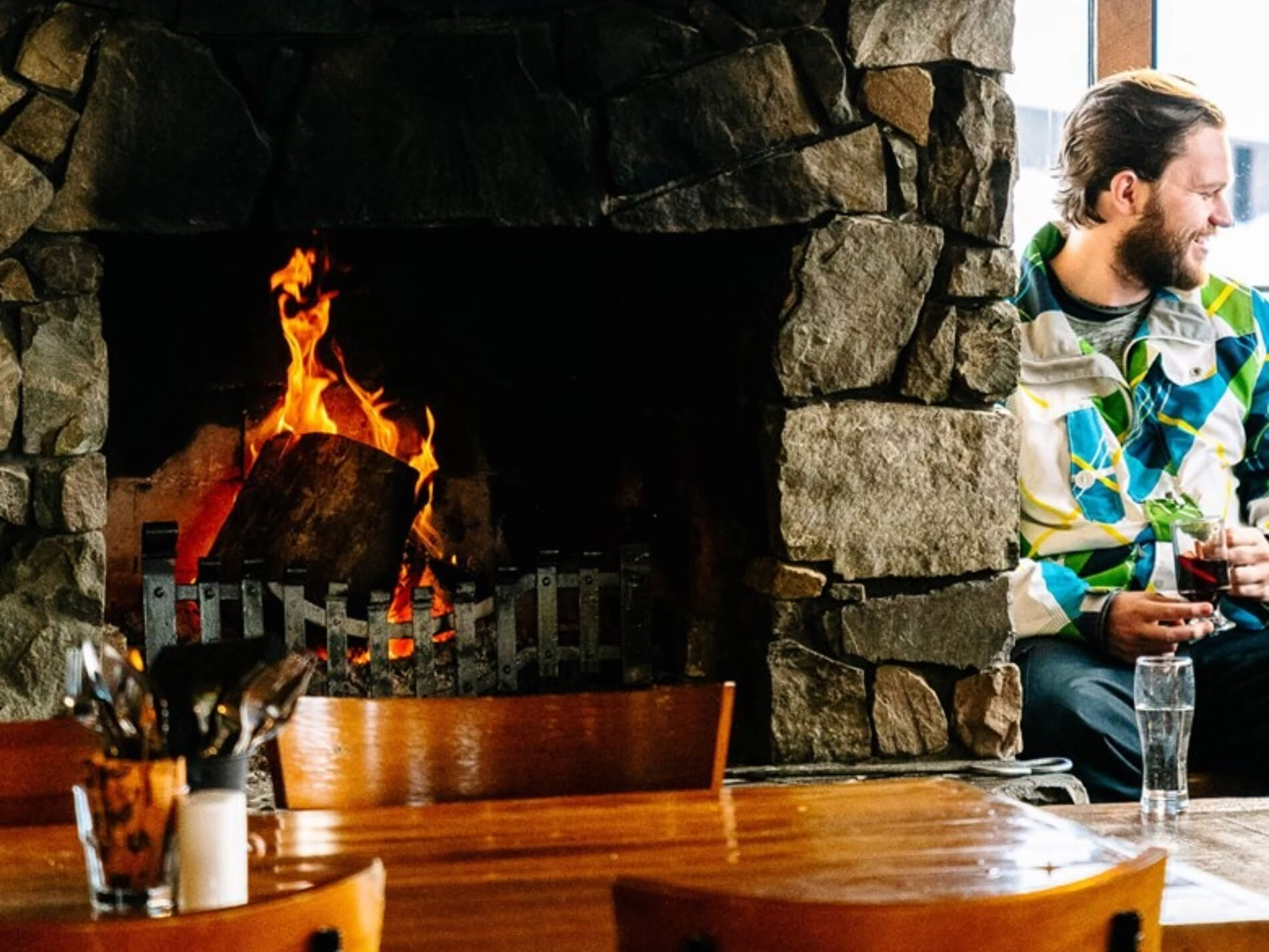 Man in colorful jacket sitting by a stone fireplace with a glass of a dark beverage, wooden table with utensils in front.