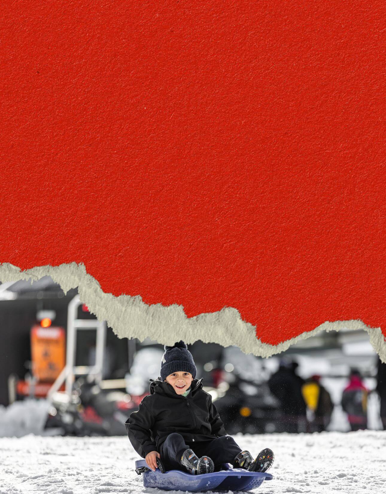 Smiling young boy in winter clothes sledding down a snowy hill on a blue sled.