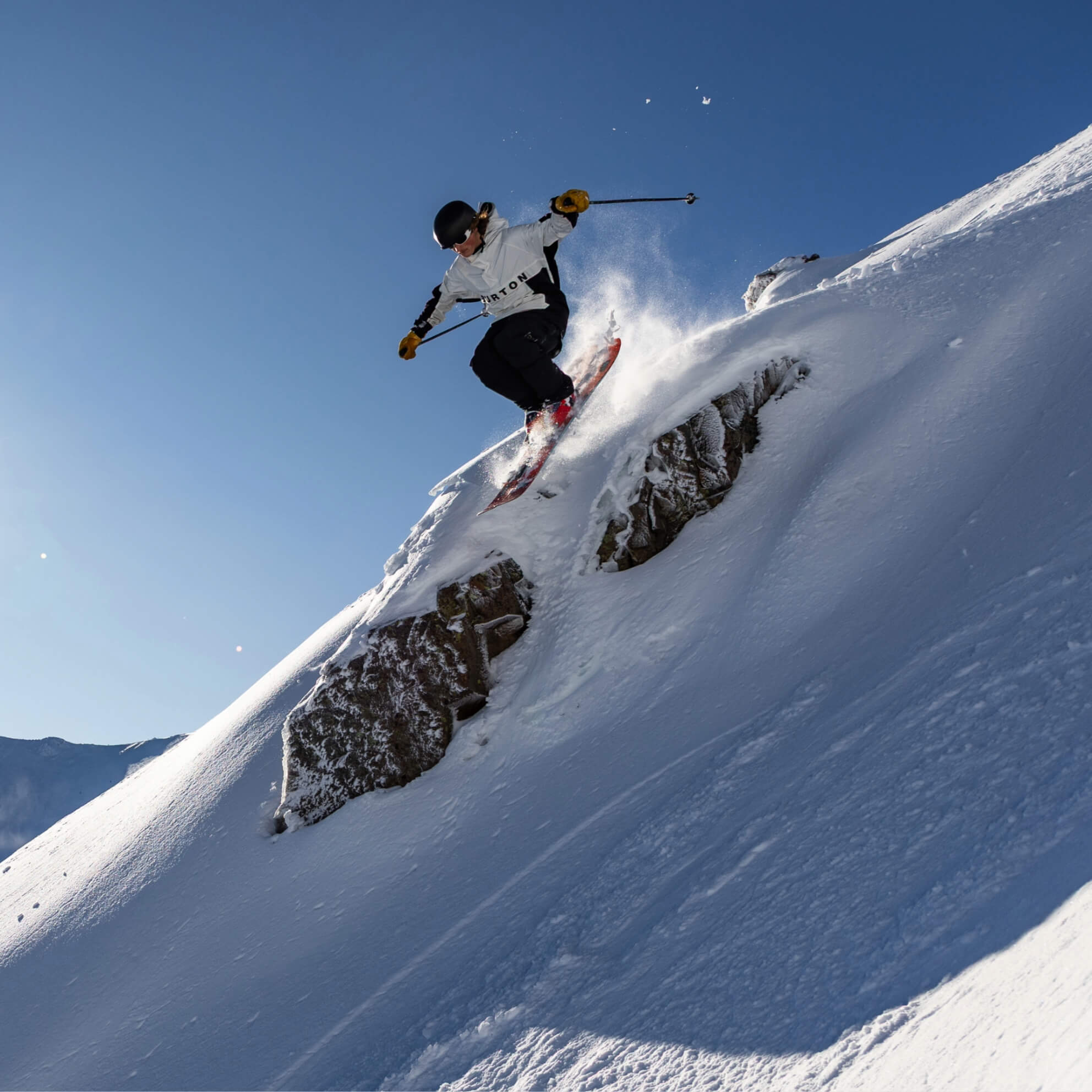 Skier wearing a helmet and white jacket jumping off a rocky snow-covered slope against a clear blue sky.