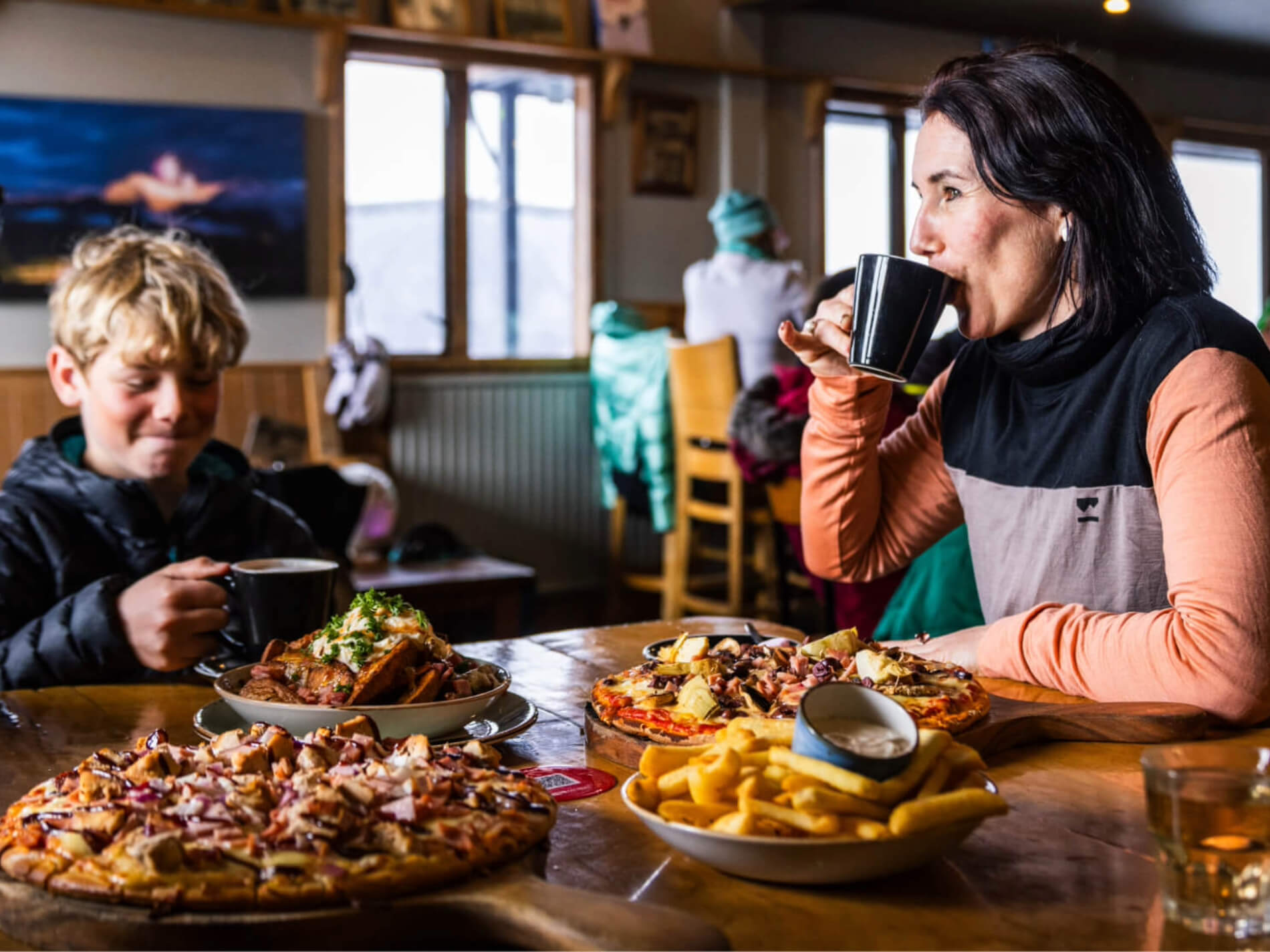 A woman and a boy sitting at a wooden table in a restaurant, drinking from black mugs with plates of pizza, fries, and a loaded dish in front of them.