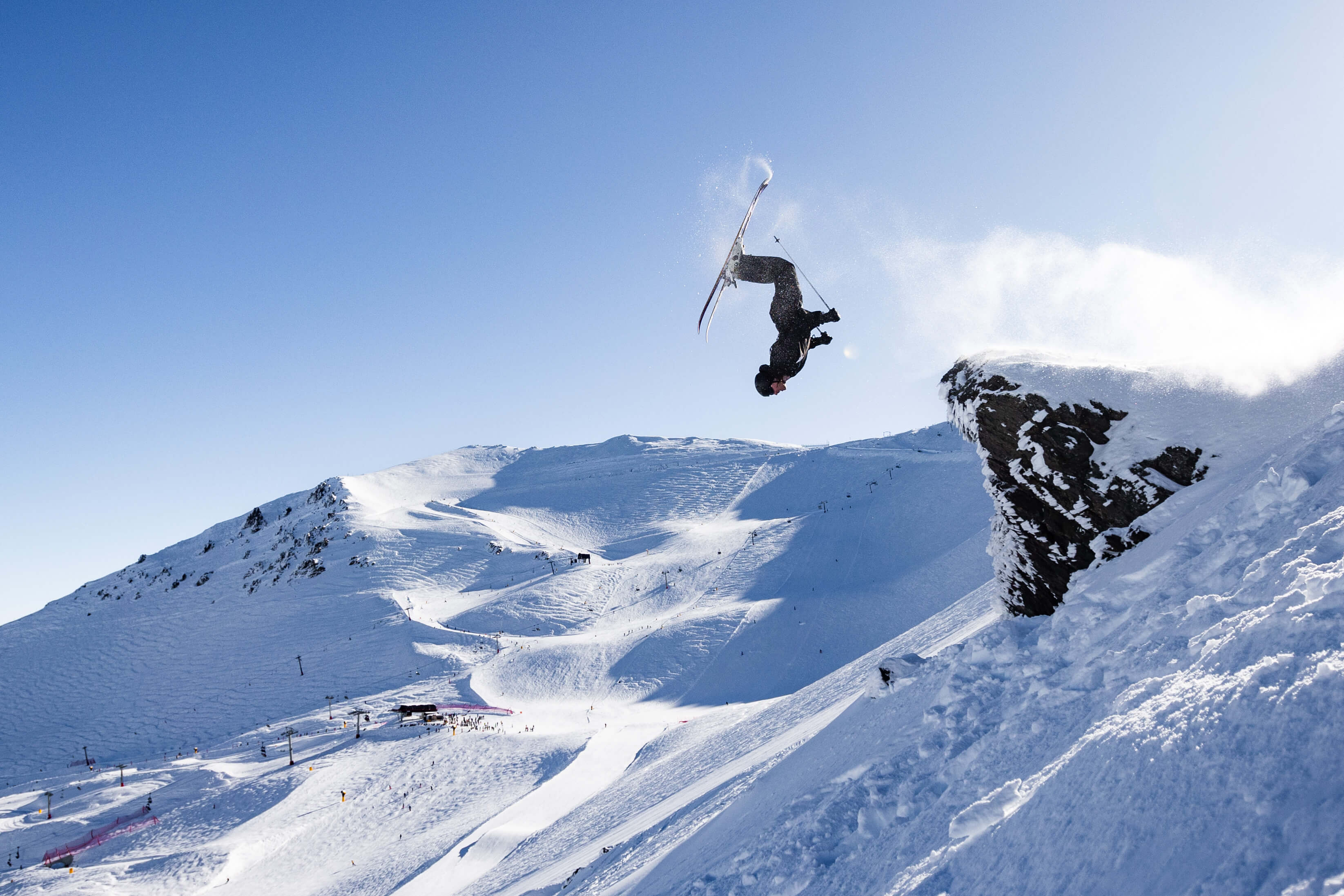 Skier performing a backflip off a snowy cliff in a mountain ski resort under clear blue sky.