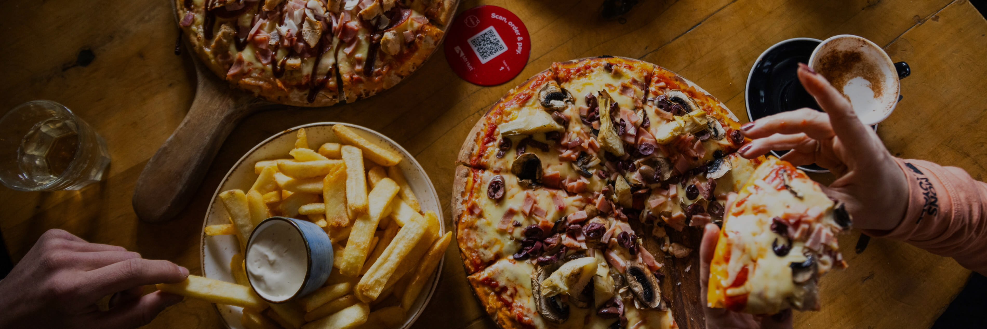 Two pizzas with toppings and a plate of thick-cut fries with dipping sauce on a wooden table, with hands reaching for food and a cup of coffee nearby.
