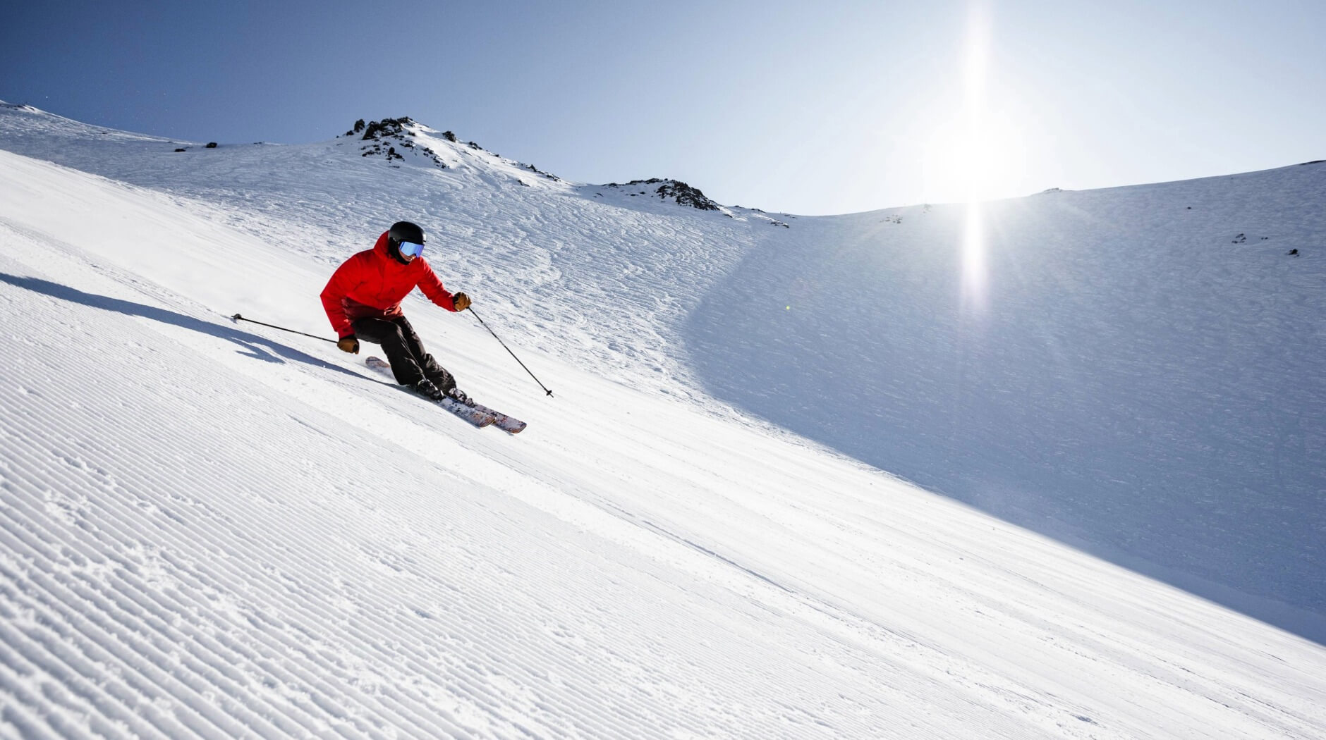 Skier in a red jacket carving turns on a groomed snowy slope with rocky mountain ridges and sun in the background.