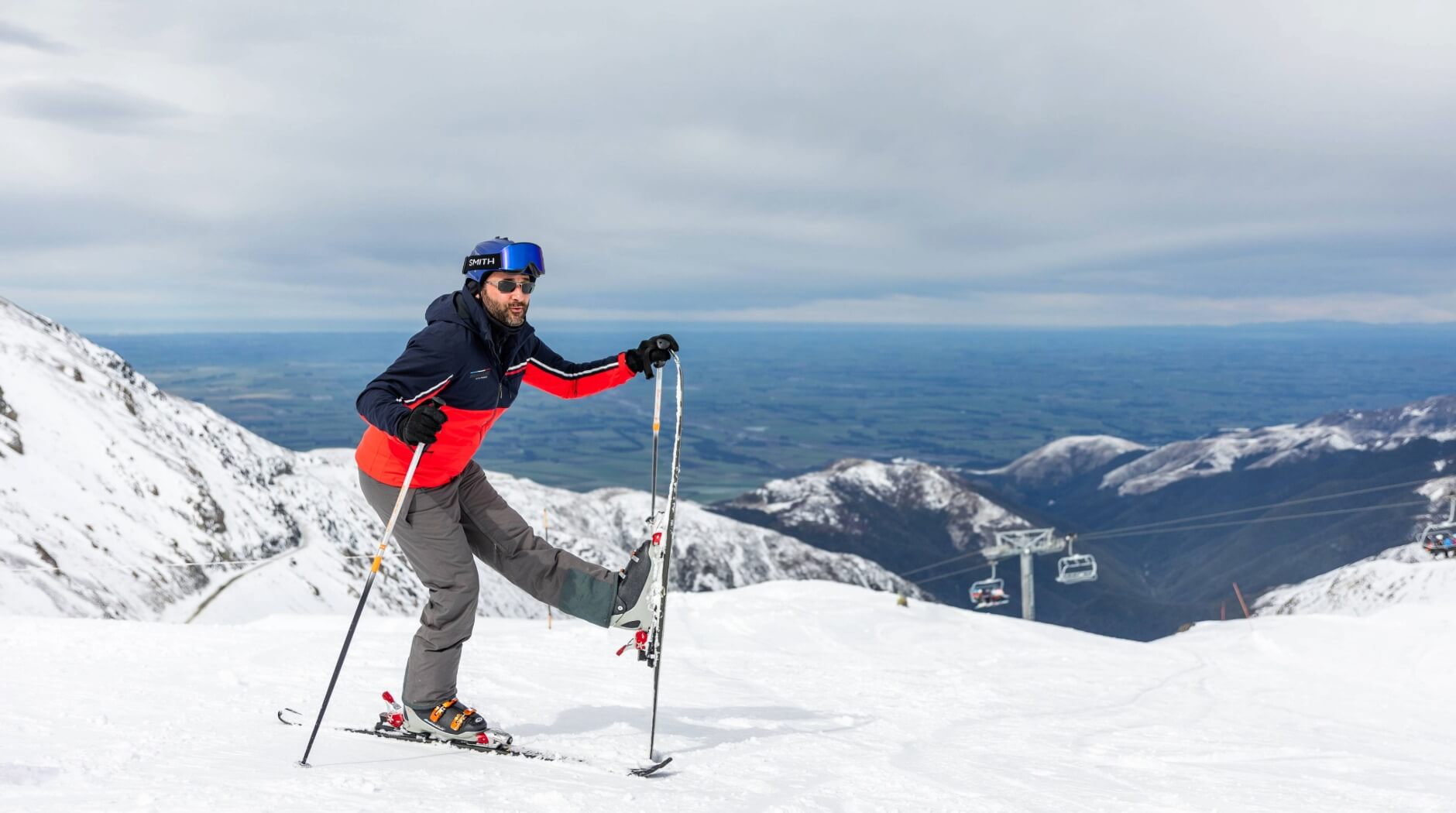 Man in red and navy jacket with blue helmet adjusting his ski on a snowy mountain slope with chairlifts and distant mountains in the background.