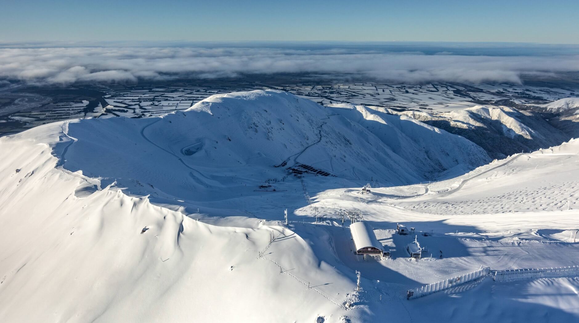 Snow-covered mountain summit overlooking ski lift infrastructure and distant farmland under a clear blue sky.