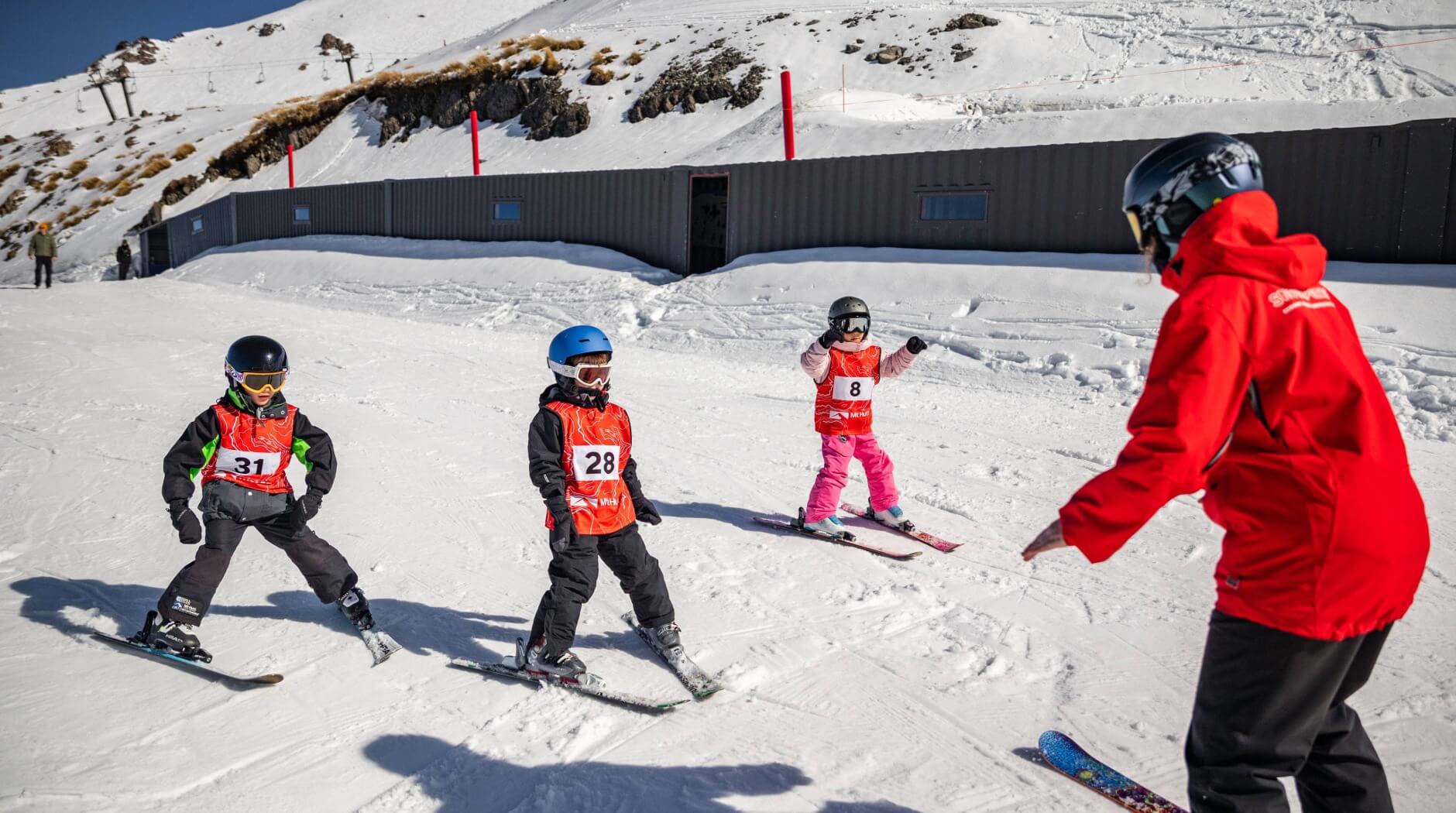 Three children wearing helmets and numbered red bibs skiing on snow while an instructor in a red jacket guides them.