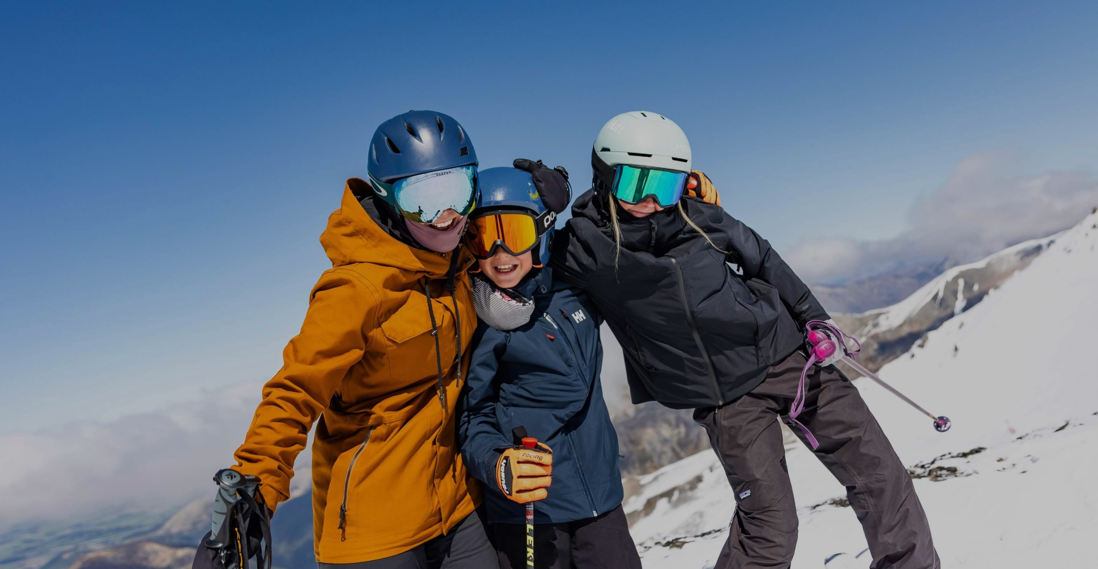 Three skiers in winter gear and helmets posing closely together on a snowy mountain under a clear blue sky.