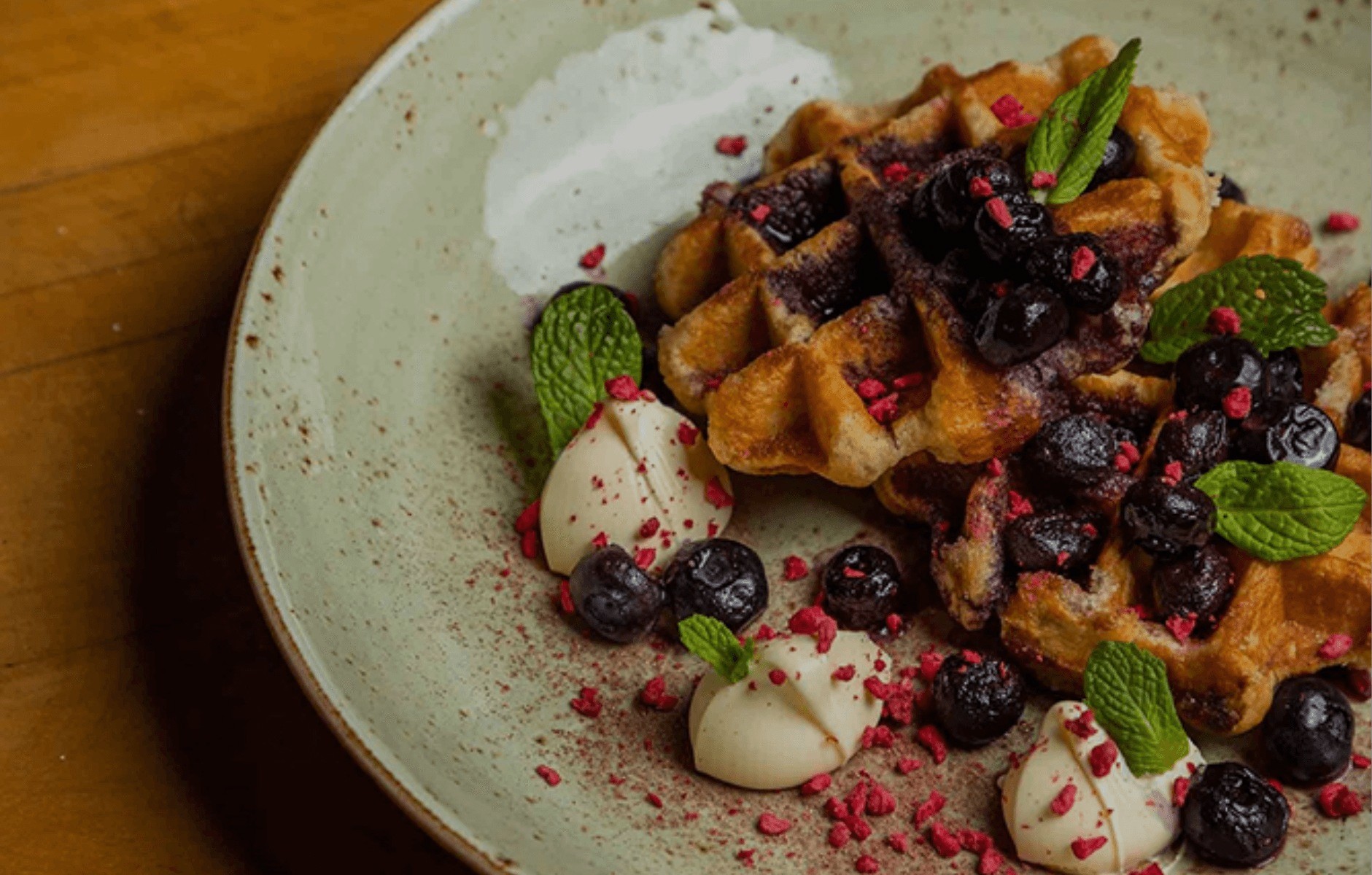 Plate with waffles topped with blueberries, garnished with mint leaves and dollops of cream sprinkled with red crumbs.
