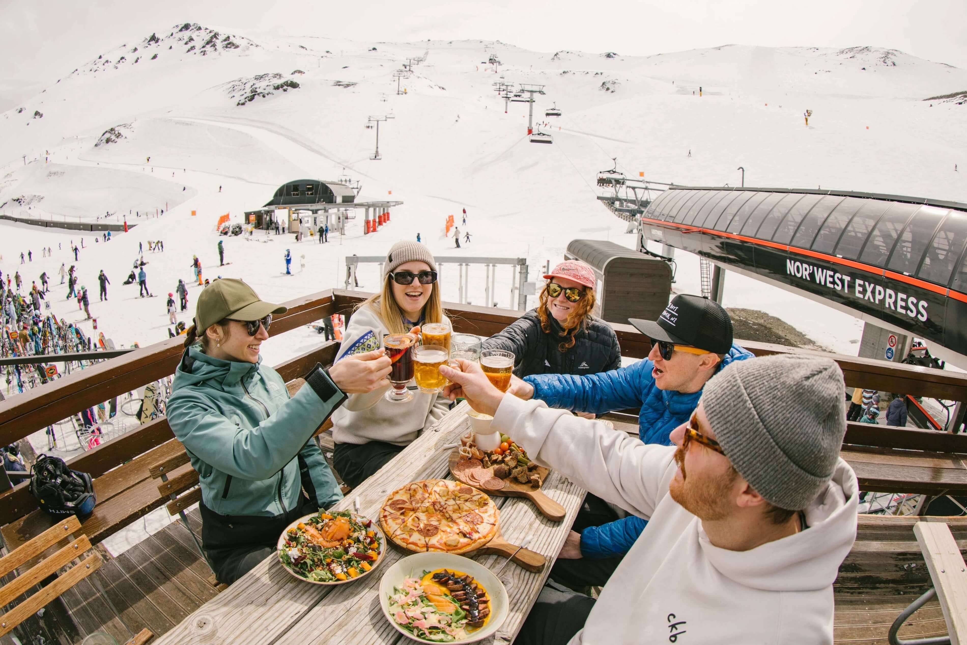 Group of five friends clinking glasses and enjoying pizza and salads on a snowy mountain deck near a ski lift.
