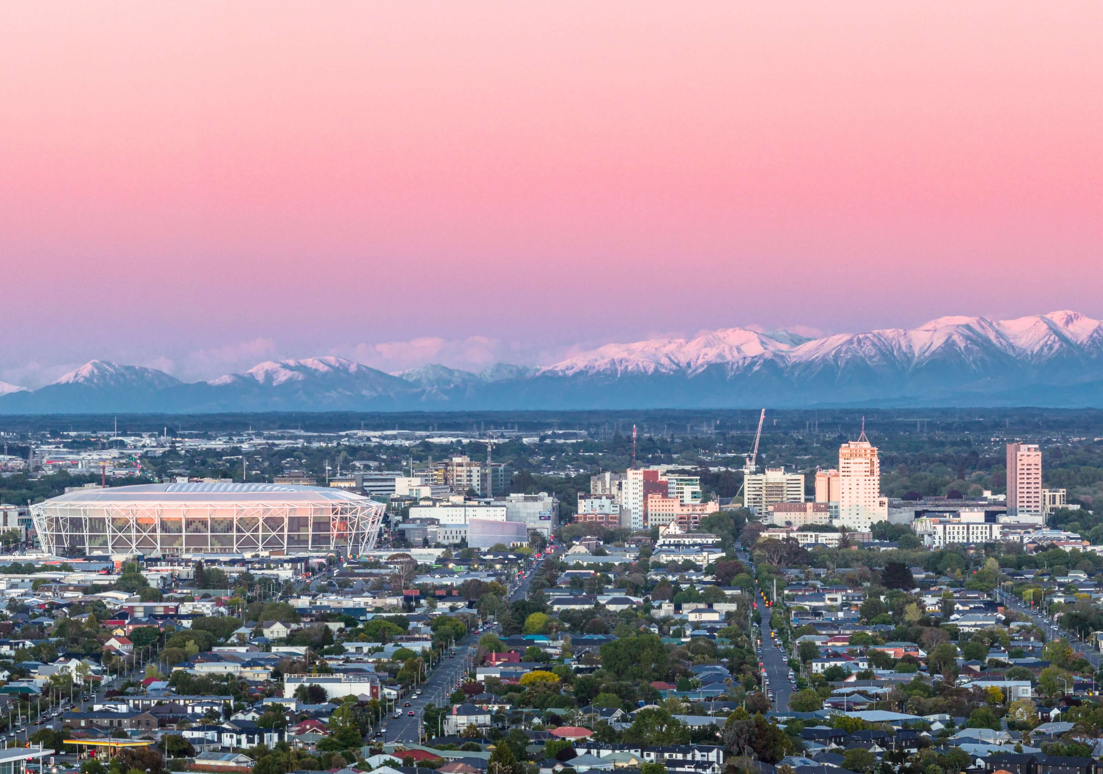 Cityscape of Christchurch with residential buildings and a stadium, set against snow-capped mountains under a pink and blue sunset sky.