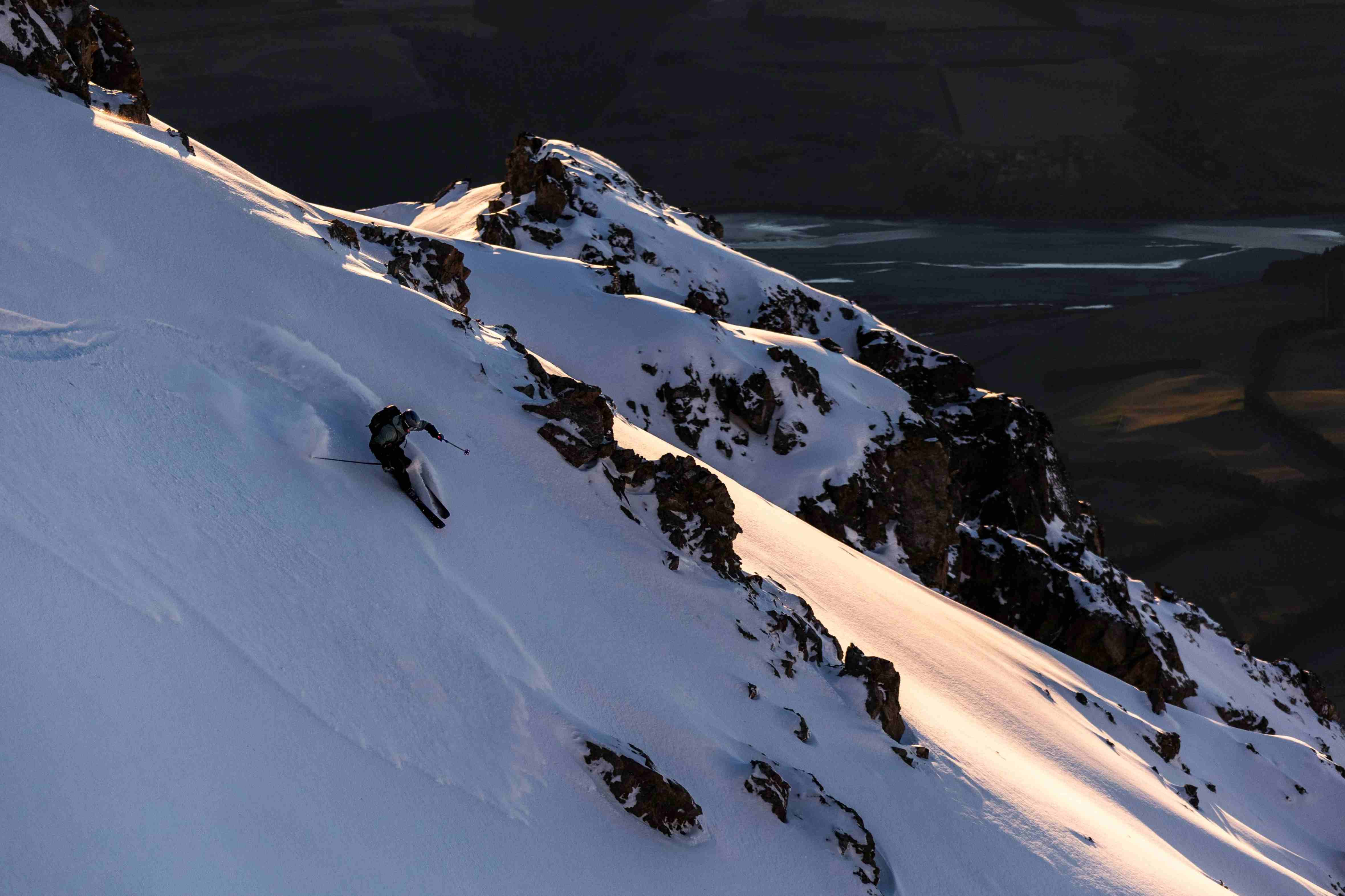 Skier making a sharp turn on a steep snow-covered mountain slope with rocky outcrops at sunset.