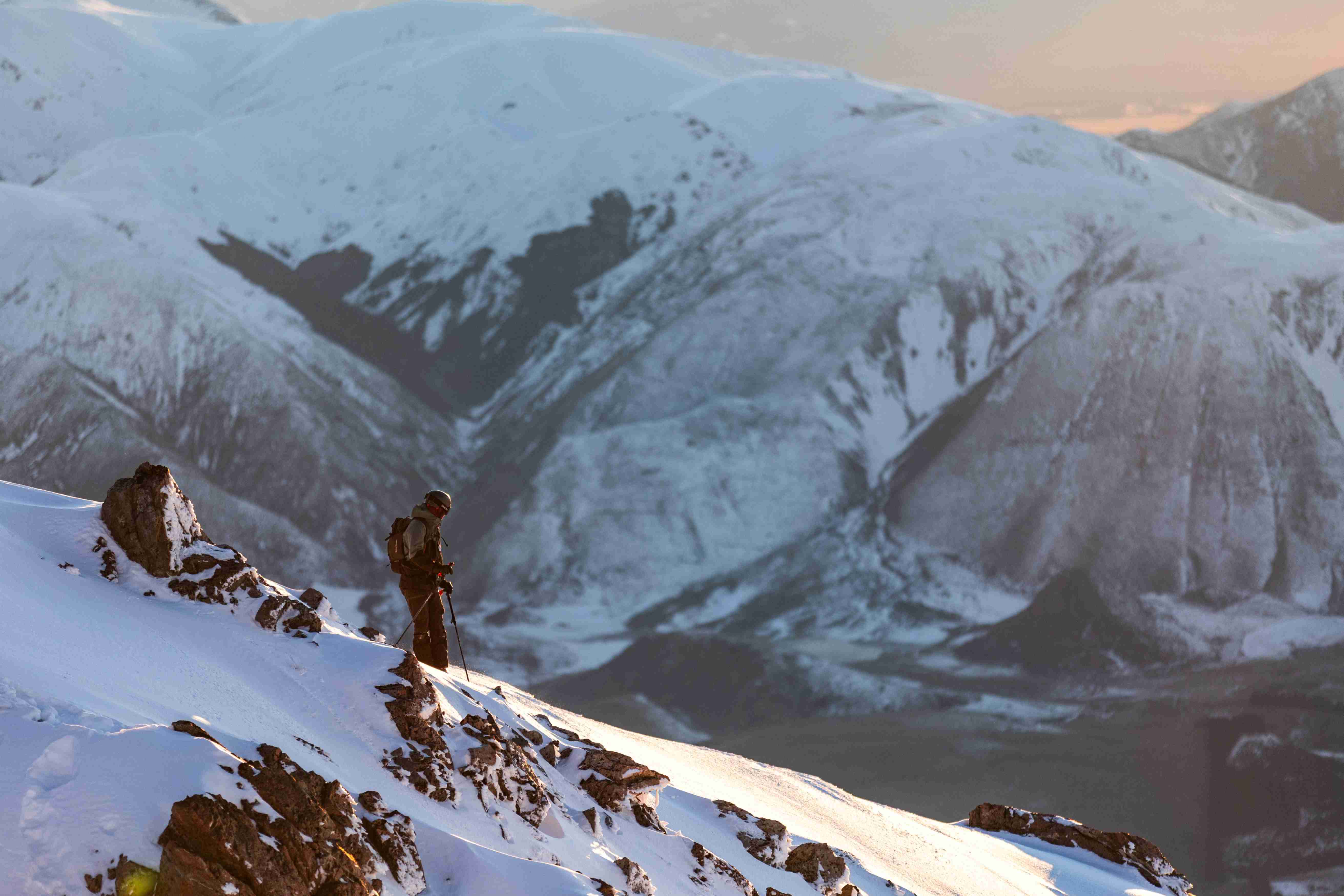 A skier standing on a snowy mountain slope with poles, overlooking snow-covered mountains in soft sunset light.