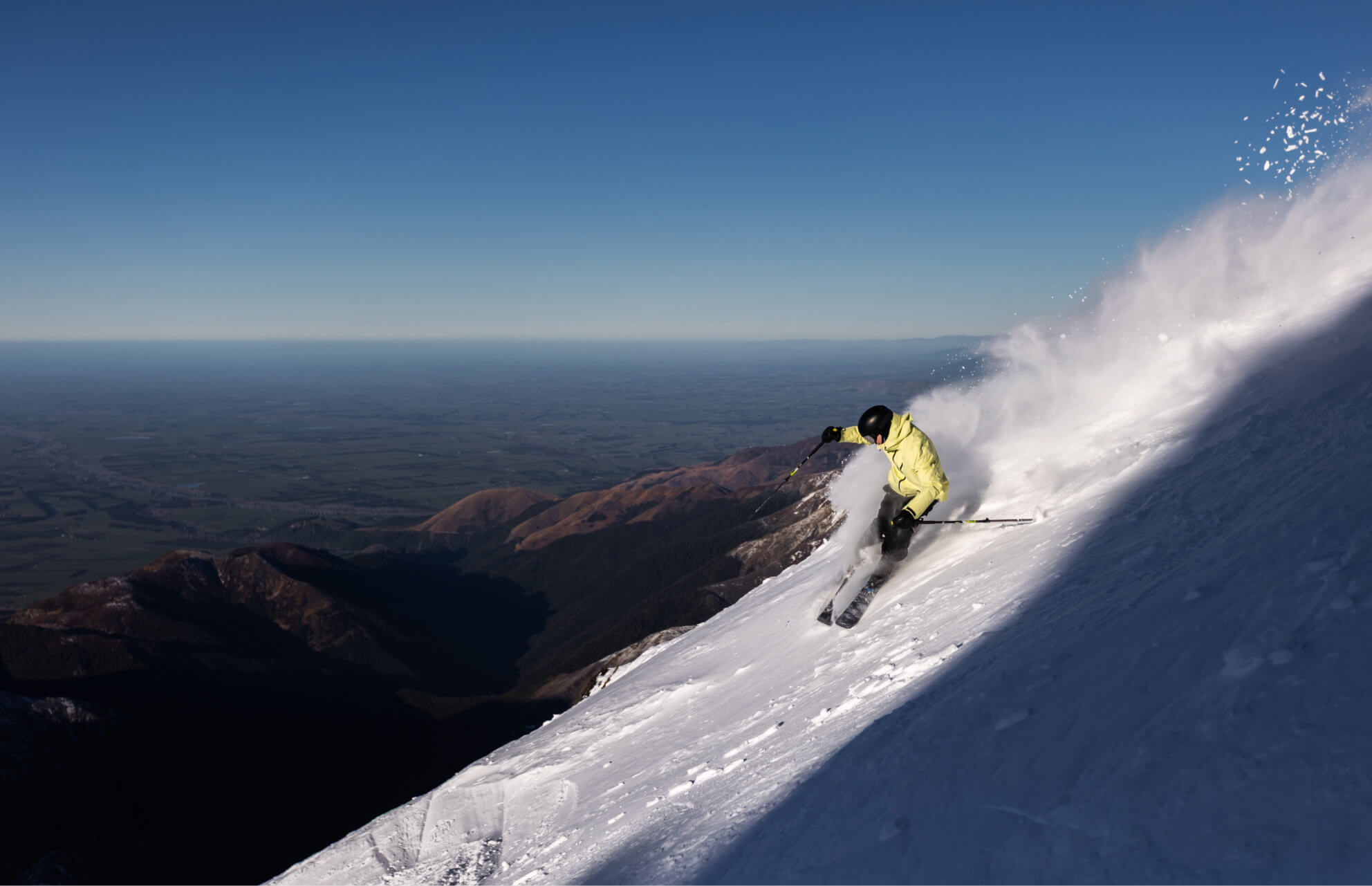 Skier in a yellow jacket skiing down a steep snowy mountain slope with snow spray against a clear blue sky.