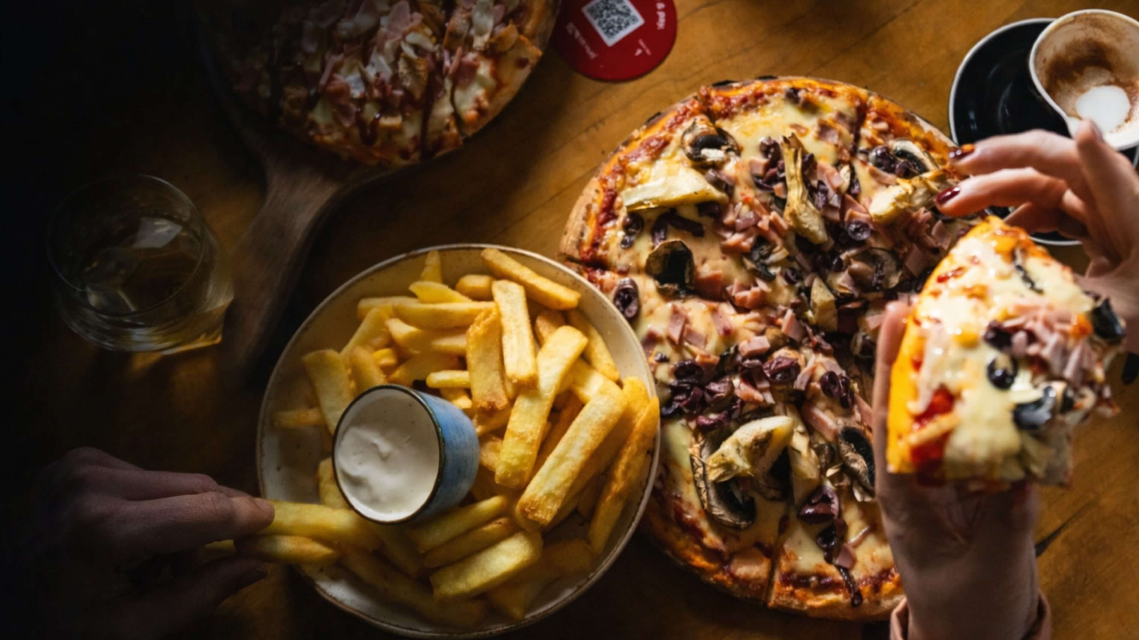 Hands holding a slice of mushroom and olive pizza next to a bowl of fries with dipping sauce on a wooden table.