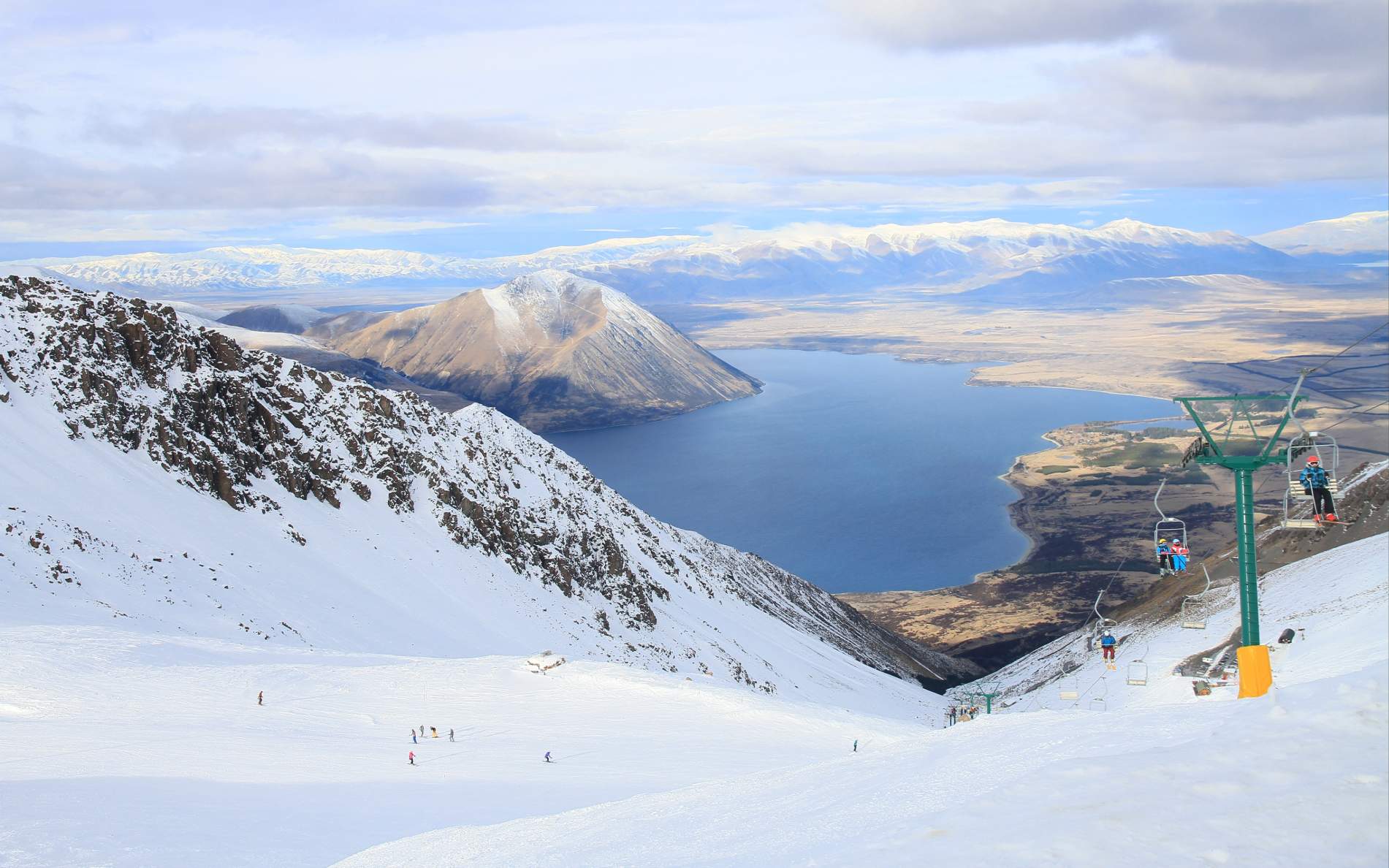 Skiers and chairlifts on snowy mountain slopes overlooking Lake Ohau and distant snow-capped mountains under a cloudy sky.