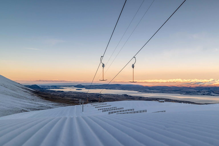 Groomed snowy ski slope with chairlift cables overhead, overlooking a lake and mountain range at sunset.