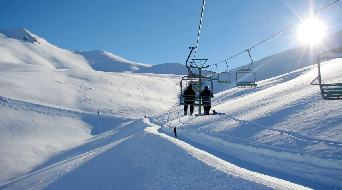 Ski chairlift carrying two people over snow-covered mountains under a clear blue sky with bright sunlight.