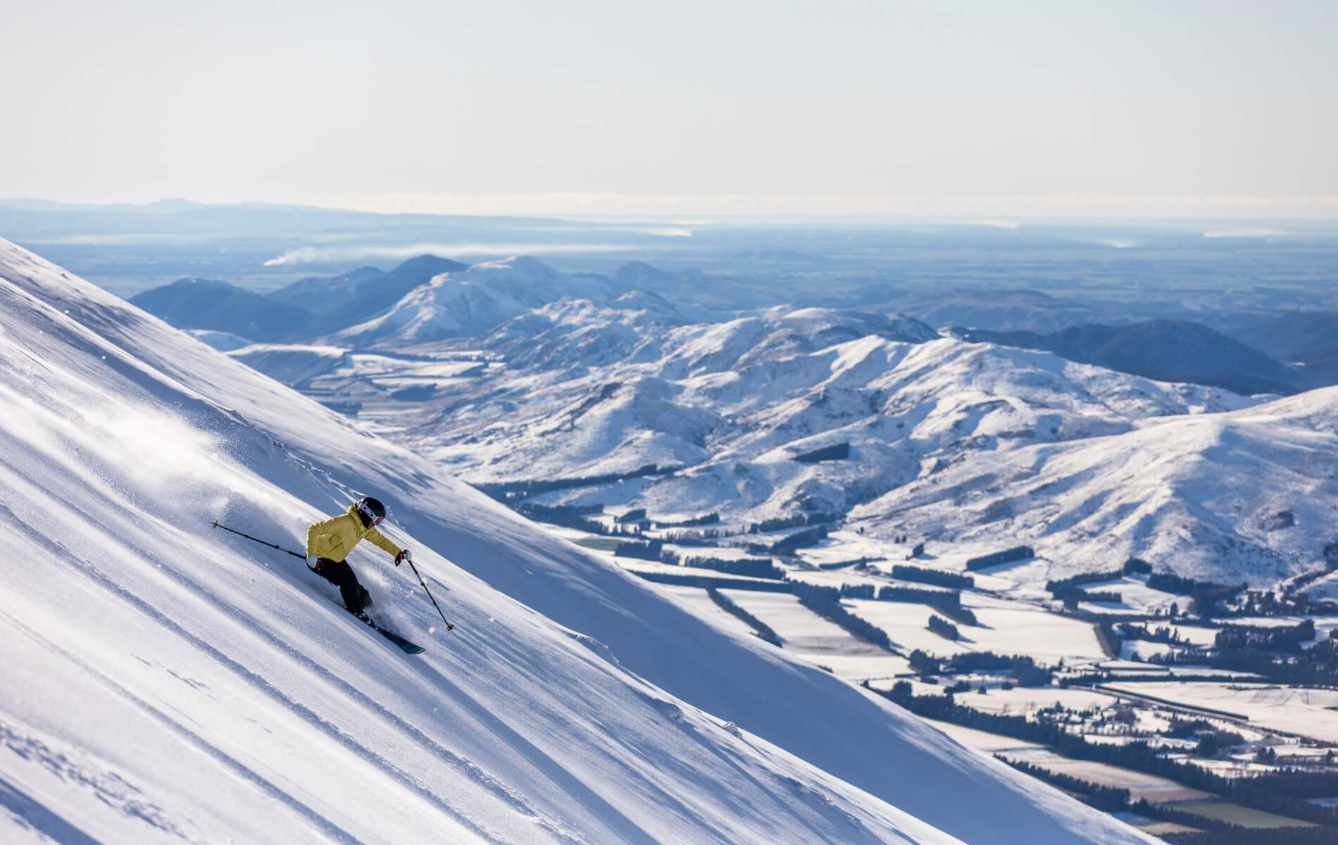 Skier in a yellow jacket skiing downhill on a snowy mountain with a vast snow-covered mountain landscape in the background.