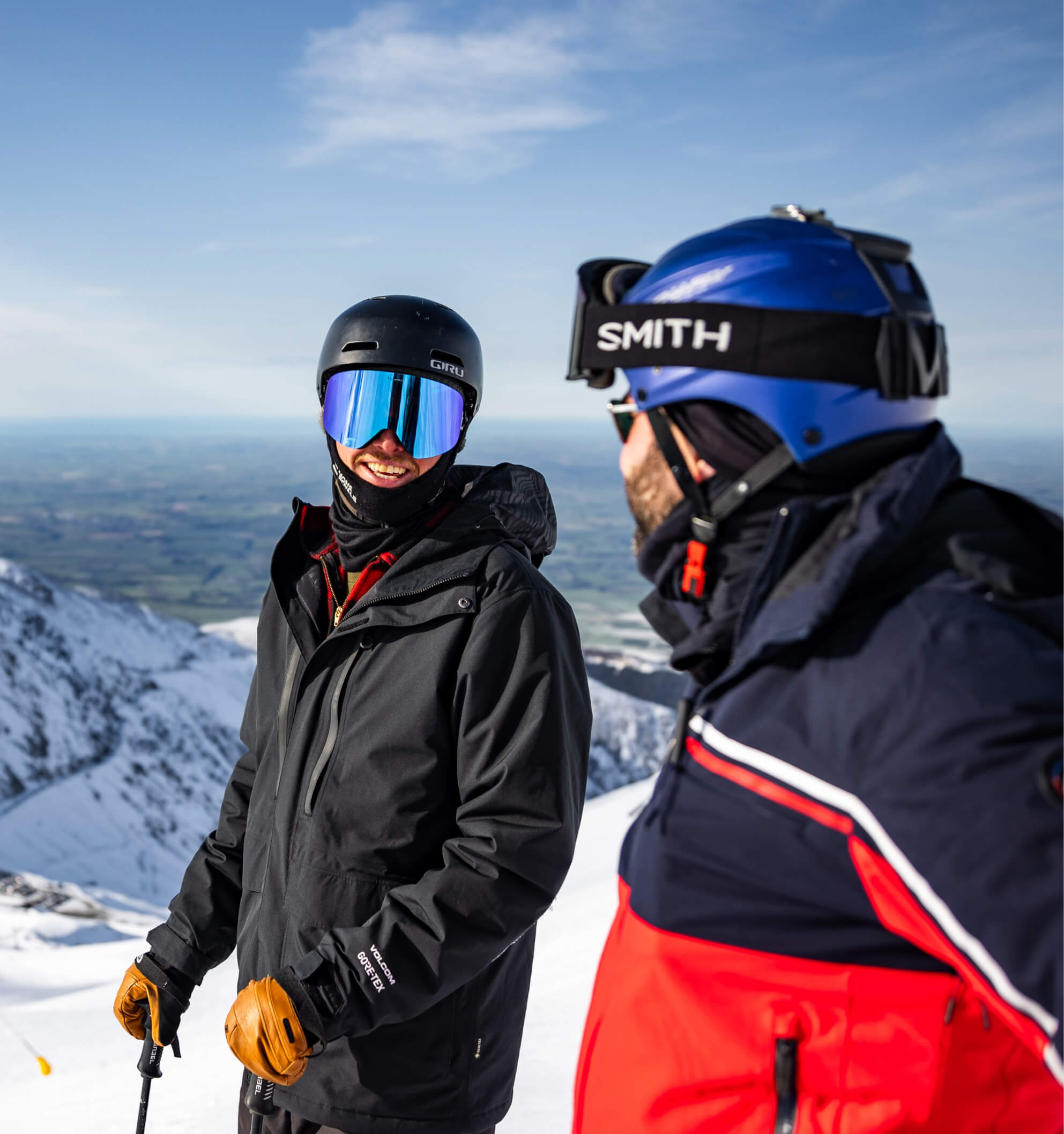 Two skiers in helmets and goggles smiling and chatting on a snowy mountain slope.