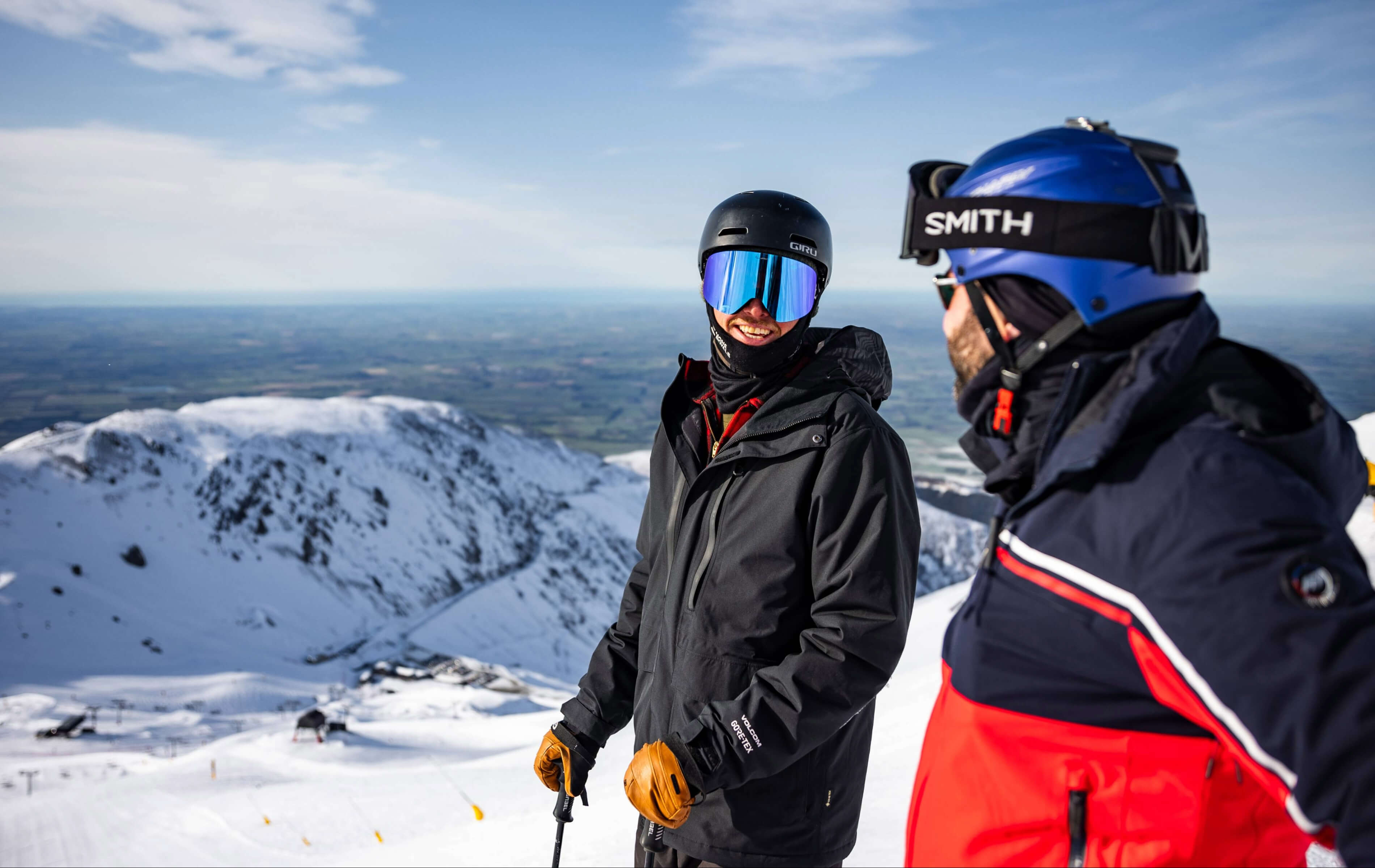 Two skiers chatting on a snowy mountain top wearing helmets and goggles with a clear sky above.