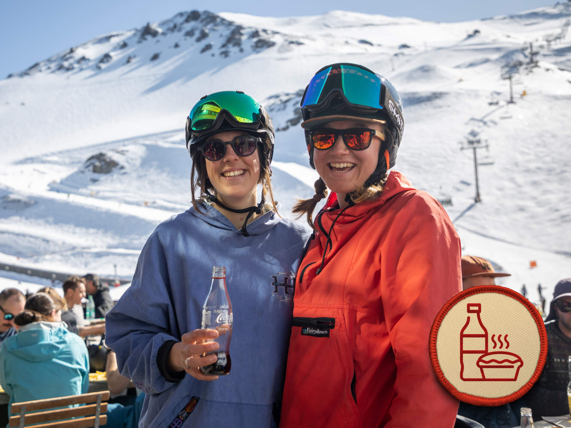 Two smiling women in ski gear with helmets and sunglasses, one holding a Coca-Cola bottle, with snow-covered ski trails behind them.